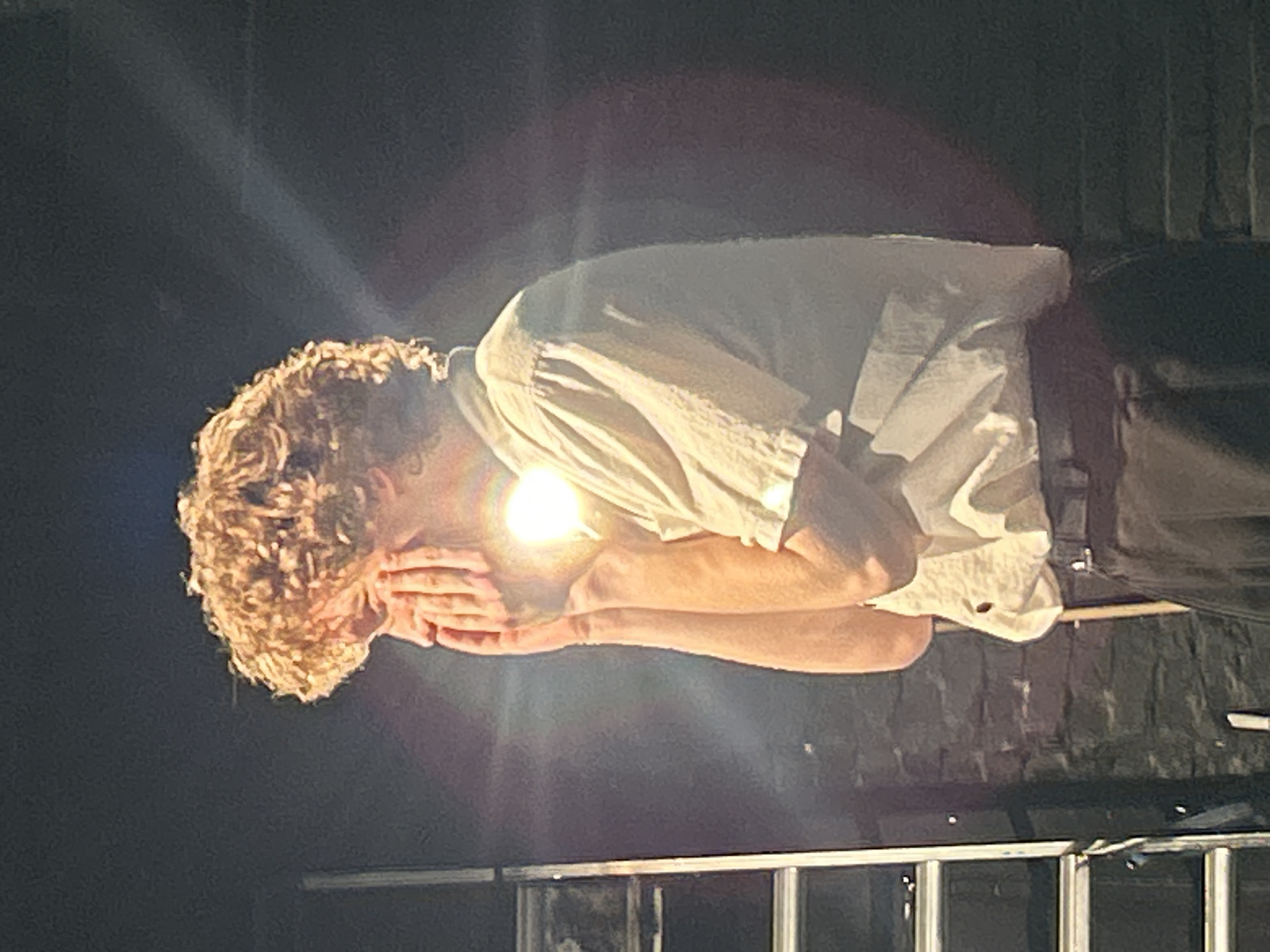 A person with curly blond hair is hunched over with their hands covering their face, illuminated by a bright light. They are wearing a loose, light-colored shirt and dark pants. The background is dark and appears to be a stage or performance area, with some scaffolding or a set piece visible at the bottom of the frame. The lighting creates a strong contrast, with the subject's face and chest brightly lit and the surrounding area in shadow. The overall mood is somber or introspective, with the subject appearing to be in distress or deep thought. There is no visible text in the image.