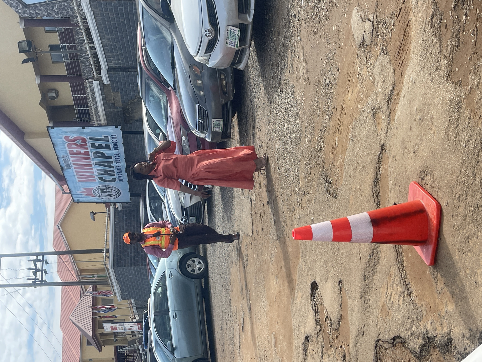 The image captures an outdoor scene at the Winners Chapel Living Faith Church in Lokogoma, Abuja, Nigeria, as indicated by a large blue sign prominently displayed on a building in the background. The sky above is partially cloudy.
The foreground and midground feature a deteriorated, unpaved ground surface characterized by numerous potholes, loose gravel, and patches of dirt, suggesting a damaged parking area. An orange and white traffic cone stands upright in the lower center foreground.
In the midground, two individuals are visible. A man is walking from left to right, wearing an orange safety vest over a pink long-sleeved shirt, dark trousers, dark shoes, sunglasses, and an orange baseball cap. He is holding a small dark object in his right hand. To his right, a woman is walking from right to left. She is dressed in a long, coral-colored dress and light-colored high heels, with her left hand raised towards her ear, possibly indicating a phone call or adjustment of an earpiece.
Numerous vehicles are parked in rows across the midground and background. Notable vehicles include a light blue sedan on the left, a dark grey Toyota Corolla with the Nigerian license plate "ABUJA YAB 302AX," and a silver Toyota Corolla with the Nigerian license plate "ABUJA ABC 207FF."
In the background, behind the vehicles, stand multiple buildings, including the church complex, featuring reddish-brown roofs and light-colored walls. A dark stone-patterned wall supports a railing in front of one building, where an outdoor air conditioning unit is also visible. Overhead, utility poles with power lines traverse the scene. Several flags are partially visible near a building on the far left.