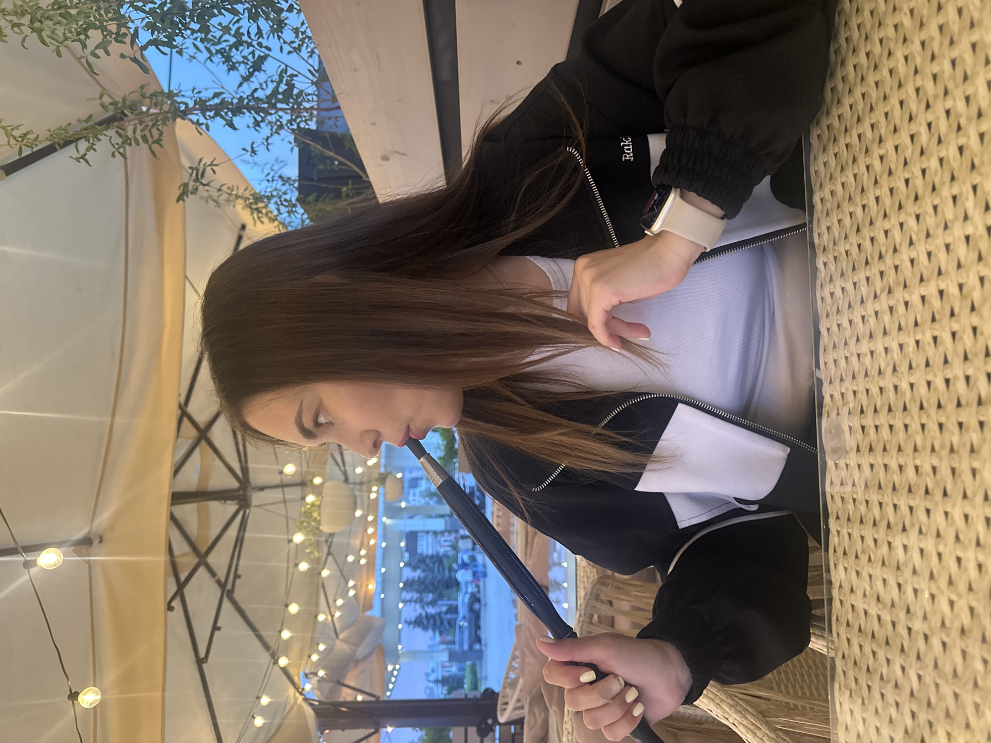 A woman is sitting at a table under a covered outdoor seating area, possibly a cafe or restaurant, with strings of lights above. She is holding a dark-colored hookah hose to her mouth, and appears to be smoking. She is wearing a black and white jacket over a white shirt and a smartwatch on her left wrist. The table in front of her has a woven surface. In the background, there is a view of trees, buildings, and parked cars, suggesting an urban setting. The overall lighting is dim, likely evening or dusk. The location is identified as Perm, Russia.