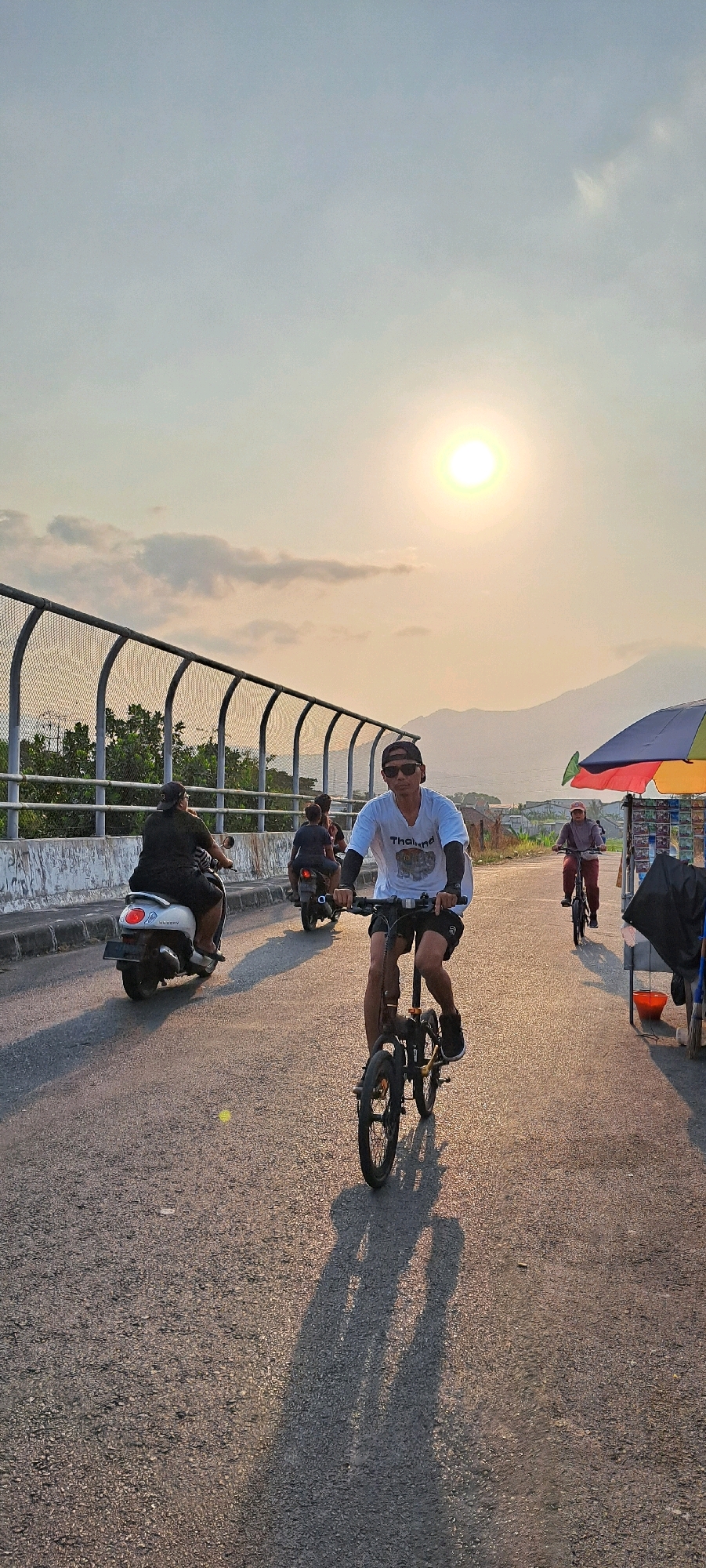 A man is riding a foldable bicycle on a road in Kebon Waris, Indonesia, during sunset. The man is wearing a white t-shirt with the word "Thailand" printed on it, black shorts, sunglasses, and a black baseball cap. He is wearing black arm sleeves and black athletic shoes. The bicycle is black and appears to be an electric bicycle. The sun is low in the sky, casting long shadows across the road.
In the background, there are several other people and vehicles on the road. To the left, a person is riding a white scooter. Behind the scooter, another person is riding a motorcycle. Further down the road, a person is riding a bicycle. On the right side of the road, there is a stall with an umbrella and various items displayed. The road is paved and appears to be a two-lane road. There is a metal fence along the side of the road, with trees and foliage behind it. In the far distance, mountains are visible. The sky is a pale blue with some clouds, and the sun is a bright orange orb. The overall lighting suggests it is late afternoon or early evening.