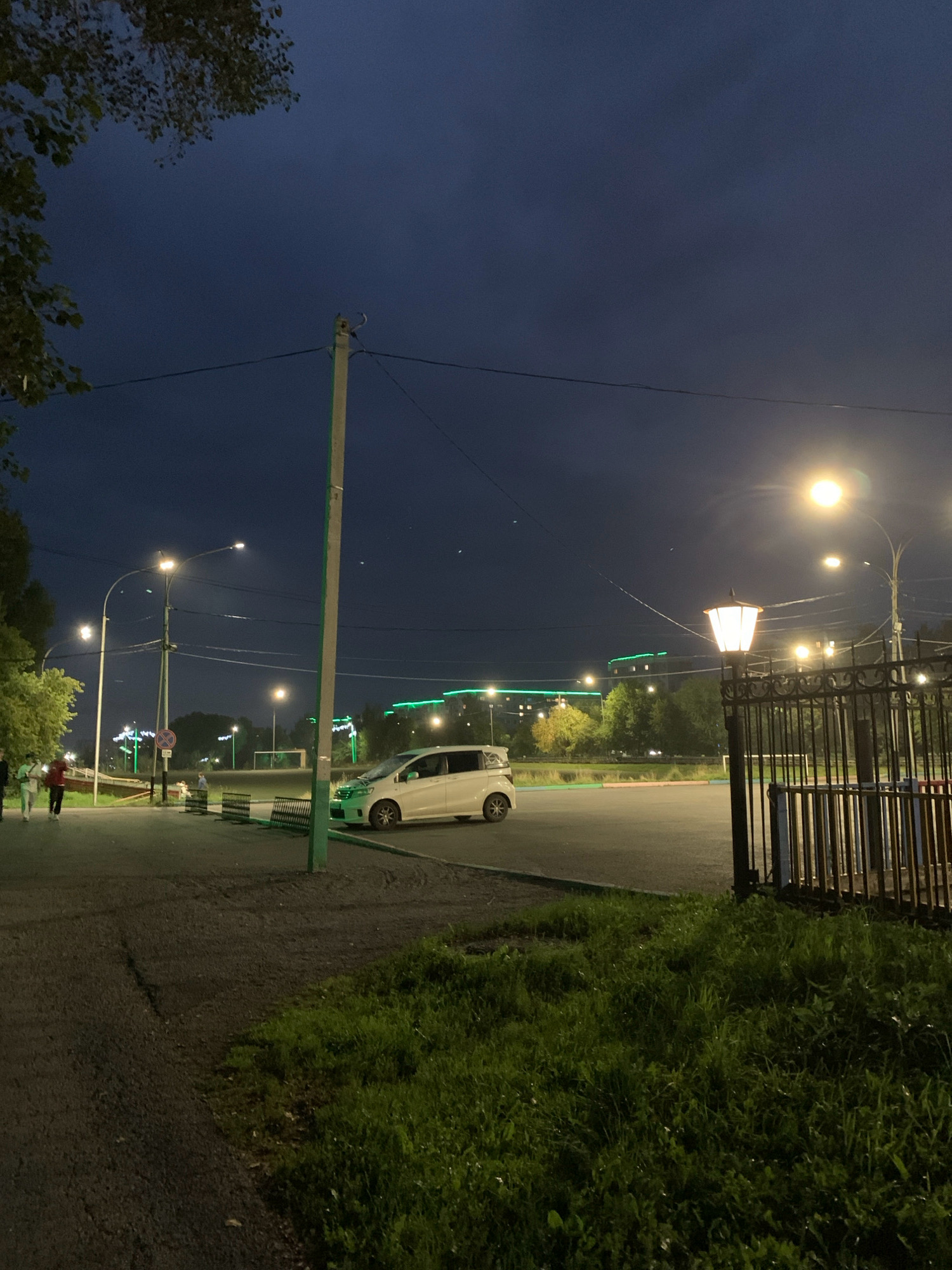 The image depicts a street scene at dusk or night in Prokopyevsk, Russia. A white minivan is parked on the right side of the frame, facing left. Several streetlights illuminate the area, casting a warm glow. In the foreground, a person in a red jacket walks on the sidewalk, followed by two individuals in white. A dark metal fence with decorative elements is visible on the right, bordering a grassy area. In the background, buildings with green lights are scattered across the horizon, suggesting an urban environment. The sky is a deep blue, characteristic of twilight or night.