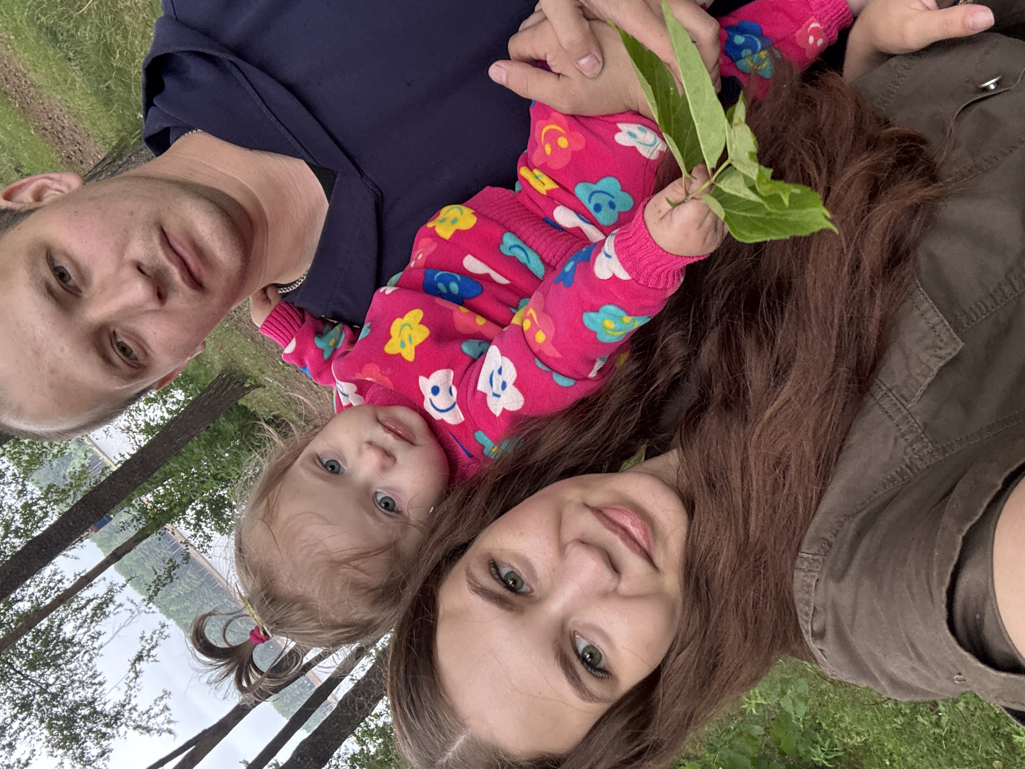 This is a selfie taken outdoors, likely in a park or natural setting, with a body of water and trees in the background. The image is oriented sideways.
The subjects are a family of three: an adult male, an adult female, and a young child. The male subject is on the left side of the frame, looking towards the camera with his head tilted. The adult female is on the right, also looking at the camera. Between them, a young child is held, also looking at the camera. The child is wearing a pink onesie with a floral and smiley face pattern. The child is holding a few green leaves. The adult male is wearing a dark blue top, and the adult female is wearing a green jacket.
The scene appears to be a casual outdoor activity, possibly a family outing or a picnic, given the relaxed poses and the natural environment. The lighting suggests it could be daytime, with natural light illuminating the subjects' faces. The emotions conveyed are generally positive and loving, with the subjects smiling or looking content.
Notable details include the child's bright pink outfit with cheerful patterns, the green leaves held by the child, and the natural backdrop of trees and water. The presence of trees and a body of water, possibly a lake or river, points towards a rural or semi-rural location. The weather appears to be pleasant for outdoor activities. There is no visible text in the image.