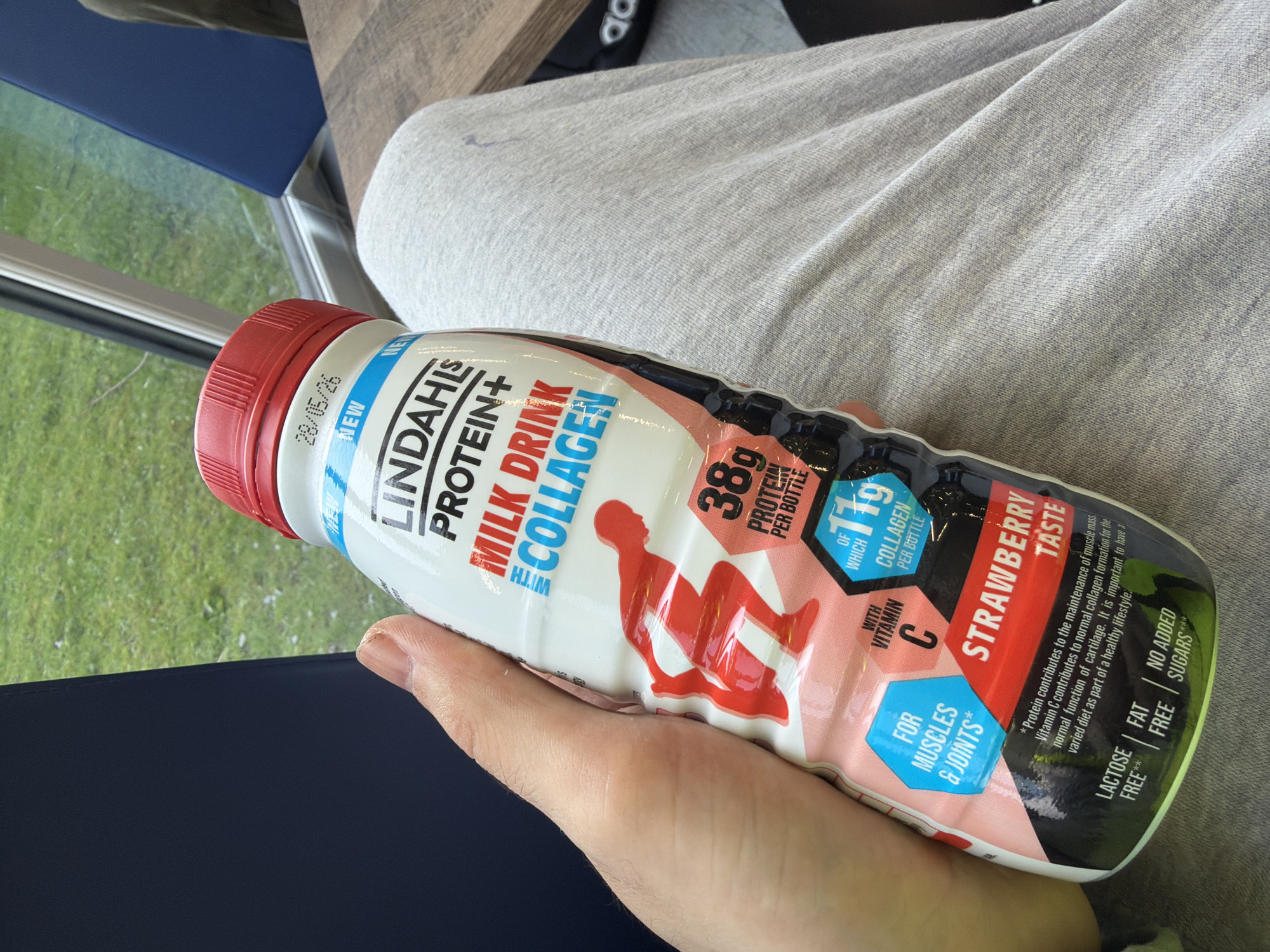 The image depicts a bottle of Lindahls Protein+ Milk Drink Collagen, strawberry flavor, held in a person's hand. The bottle is red, white, and black. The bottle claims to have 38g of protein per bottle, 11g of which is collagen. The bottle is lying on someone's lap and the person is wearing grey sweatpants. There's a blue chair and a table in the background.
The image was taken on 28/05/26.
The location is not provided in the image.