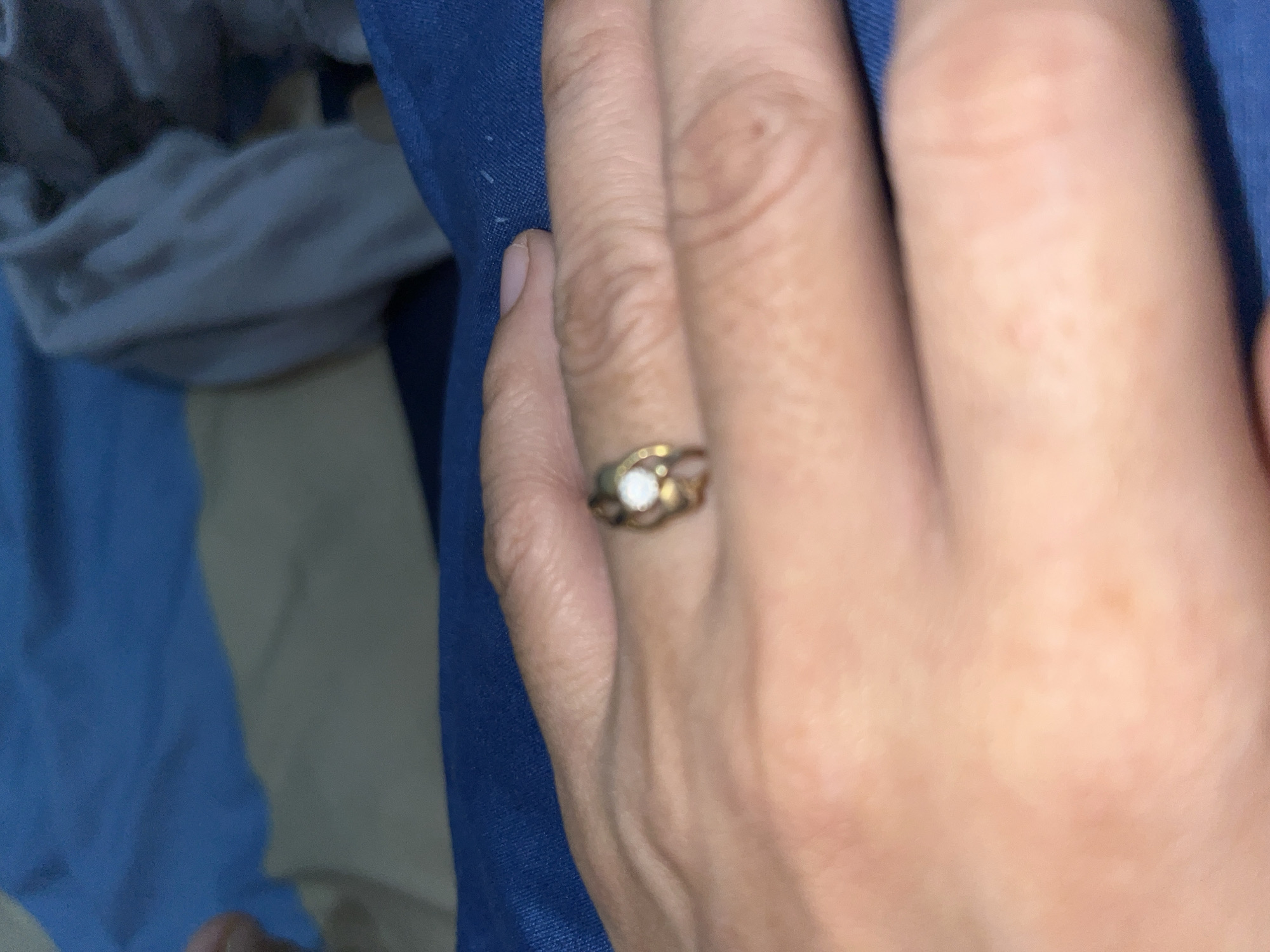 A close-up photograph captures a human hand, likely an adult's, with a medium skin tone. The hand is adorned with a ring on one finger, featuring a gold-toned band and a prominent, round, clear or white gemstone, possibly a diamond or similar stone. The band of the ring appears to have some subtle decorative detailing flanking the central stone. Visible skin texture, including creases and a small mole, is present on the fingers.
The hand is positioned against a background of textiles. A vibrant blue fabric, possibly denim or a similar material, is in sharp focus directly beneath or beside the hand. The background beyond this blue fabric is softly blurred, showing folds of light grey and additional blue fabric, suggesting an indoor or personal setting, such as a home. There is no specific activity depicted; the hand appears to be resting. The lighting in the image is warm and soft, indicating either indoor lighting or a shaded outdoor environment, but no specific time of day or weather conditions can be determined. No text is visible in the image.