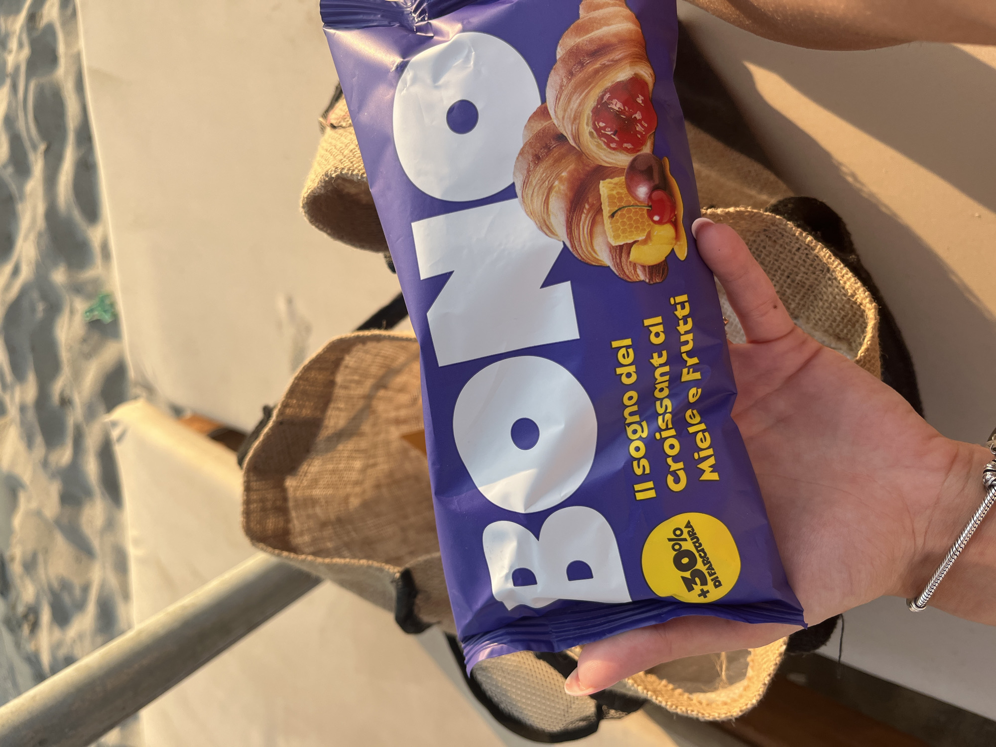A hand holds a purple package of "BONO" brand croissants with a filling of honey and fruit. The package has an image of two croissants, one with a fruit filling and the other cut open to show a honeycomb and fruit slices. A yellow circle on the package states "+30% DI FARCITURA". In the background, sand and a folded beige bag are visible, suggesting an outdoor setting, possibly a beach.