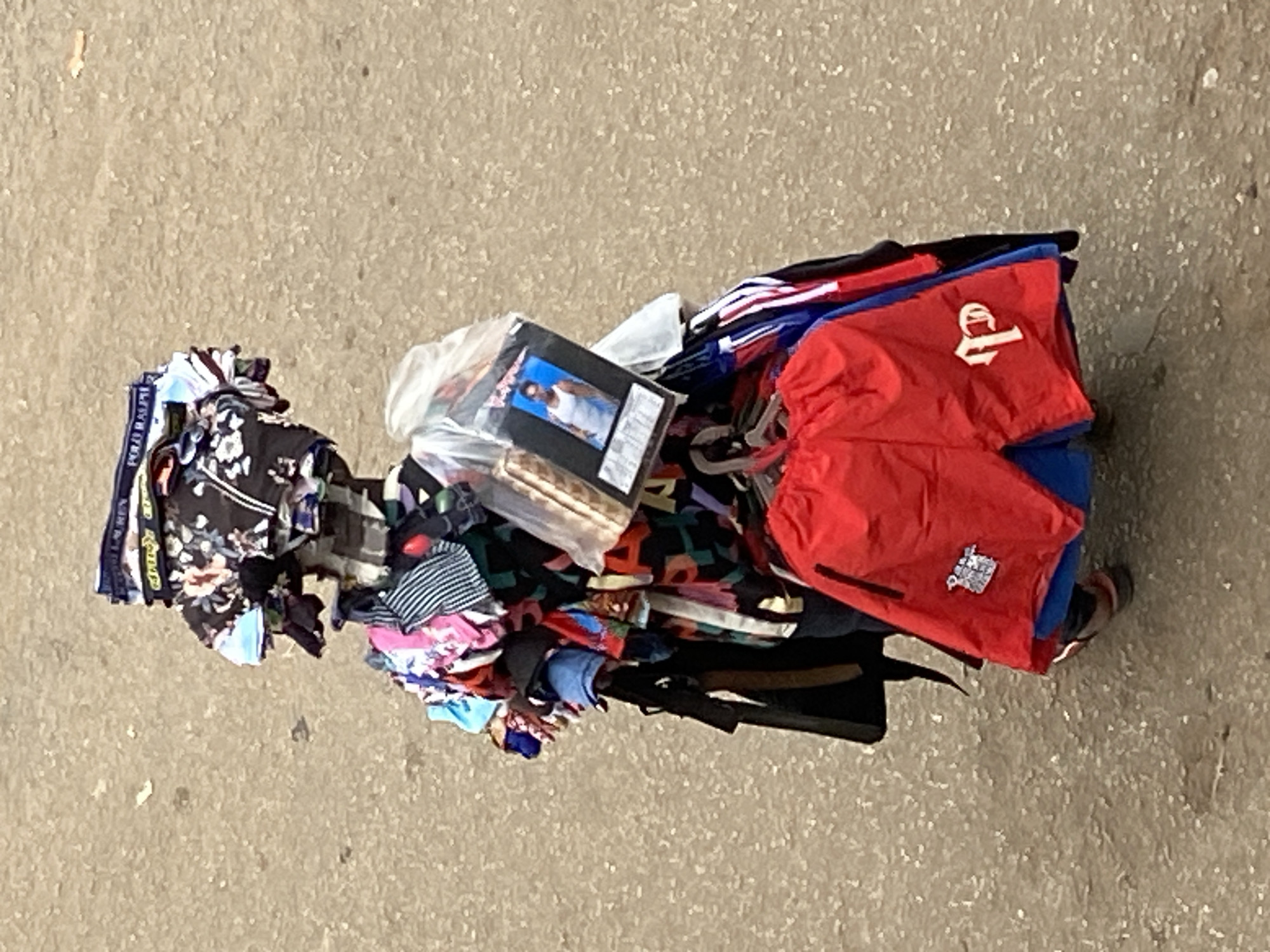 A top-down view shows a substantial, disorganized pile of various items resting on a light brown or grey textured ground surface, likely concrete or pavement. The majority of the items are articles of clothing, including patterned fabrics (such as floral and abstract designs), striped garments, and solid-colored apparel. Notably, some boxer briefs or underwear with "POLO RALPH" visible on the waistband are part of the pile. A pair of red shorts features a white stylized letter, possibly a "P" or similar initial, and a smaller white emblem on the lower leg.
Among the clothes, there's a clear plastic bag containing other objects. A distinct black-covered package, possibly a book, DVD case, or album, displays a photograph of a man in a white tank top. Partially visible beneath the main heap of items is a dark, elongated object that strongly resembles a rifle or shotgun. At the very bottom right of the pile, a foot clad in a sandal is visible, suggesting a person is standing next to or carrying these goods.
The scene appears to be set outdoors in daylight. The collection of items, particularly the diverse clothing and packaged goods, hints at the inventory of a street vendor or items being transported for sale. The presence of the visible firearm is a significant and notable detail within the context of the gathered goods.