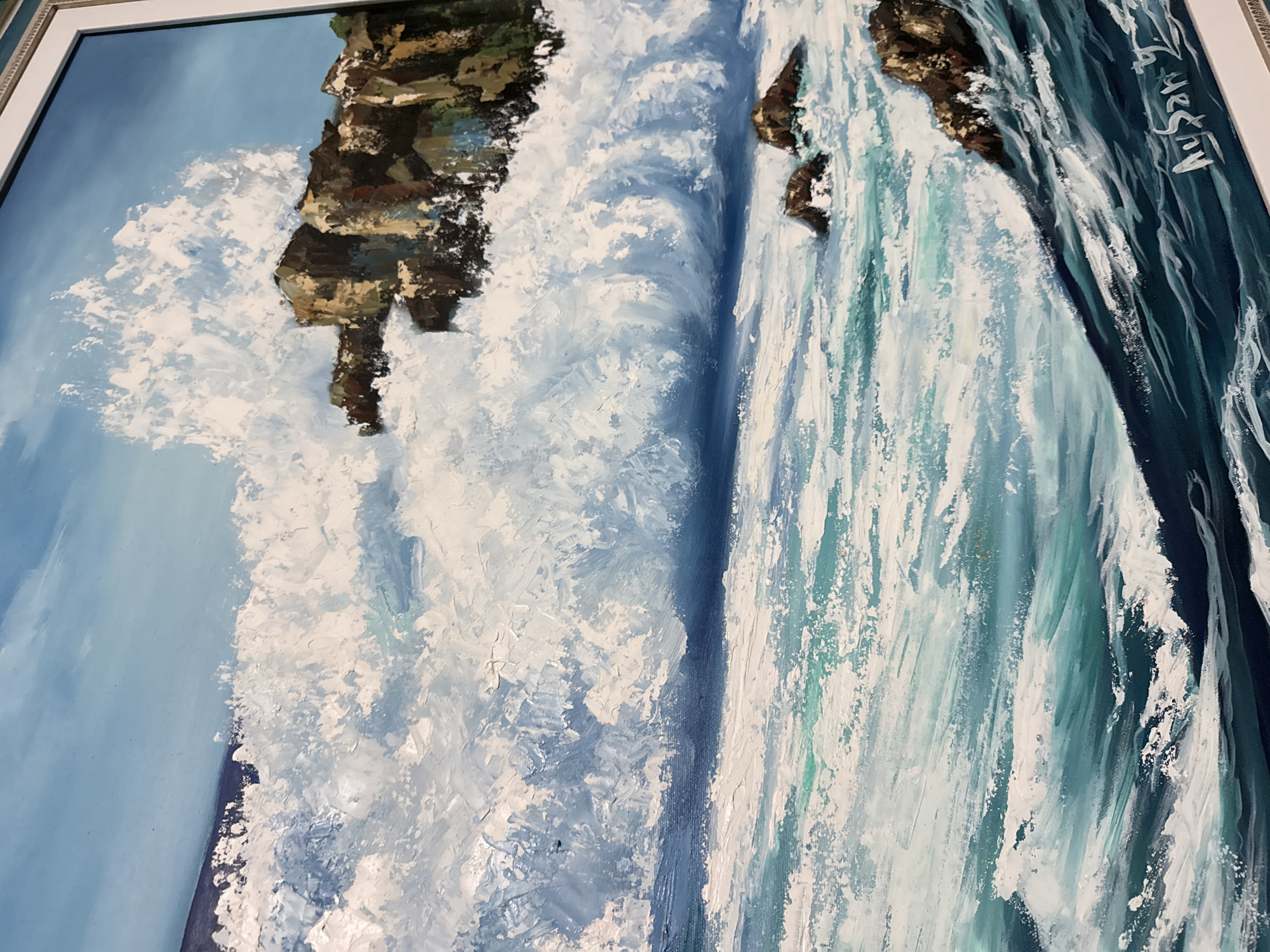 This is an oil painting on canvas depicting a dramatic seascape with crashing waves against rocky outcrops. The sky is a clear, bright blue, with textured white brushstrokes representing foam and spray. The ocean is rendered in various shades of blue and green, with white highlights indicating turbulent water and breaking waves. Several dark brown rocks are visible, some emerging from the water and others forming a larger mass against which the waves are breaking. The painting is framed in a white border with decorative molding. A signature, "AySan", is visible in the lower right corner of the painting. The scene is purely naturalistic and does not contain any identifiable elements that would suggest a specific location within Sabunchu Raion, Azerbaijan, or any other urban or suburban setting.