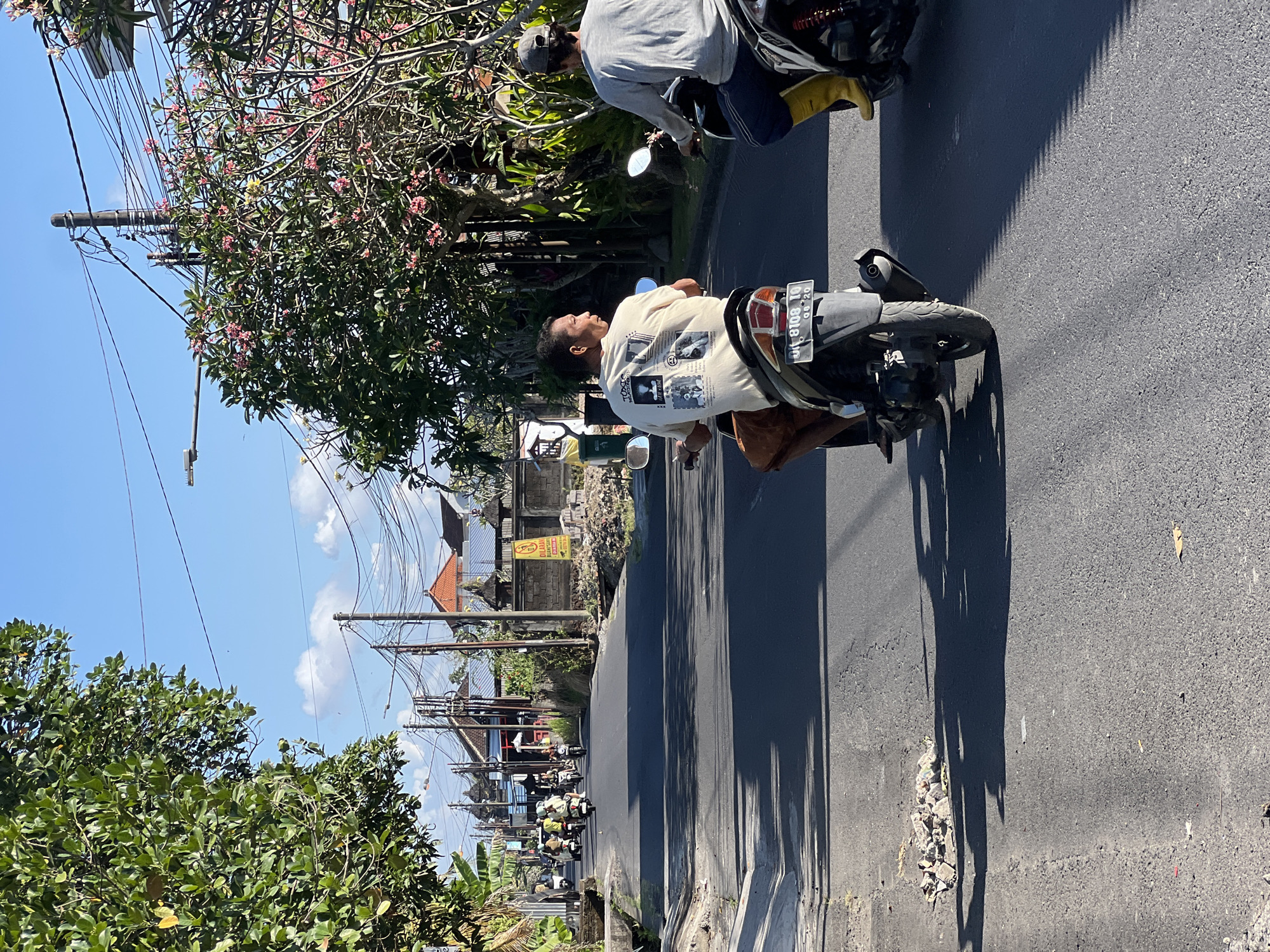 The image depicts a street scene in Denpasar, Indonesia, likely a residential or commercial area given the presence of houses and shops. The road is asphalt, with a narrow concrete drainage ditch on the left side. Several motorcycles are present, some carrying one or two people. In the foreground, two men on motorcycles are prominent. The man on the left, wearing a light-colored t-shirt with a graphic print and brown shorts, is looking to his left. The man on the right, wearing a grey long-sleeved shirt and dark pants with bright yellow boots, is looking forward. Both are riding motorcycles down the street. In the background, other motorcycles and people are visible further down the road, along with buildings, trees, and power lines overhead. The sky is blue with scattered clouds, suggesting a sunny day. The overall impression is of a typical day in a bustling Indonesian town.