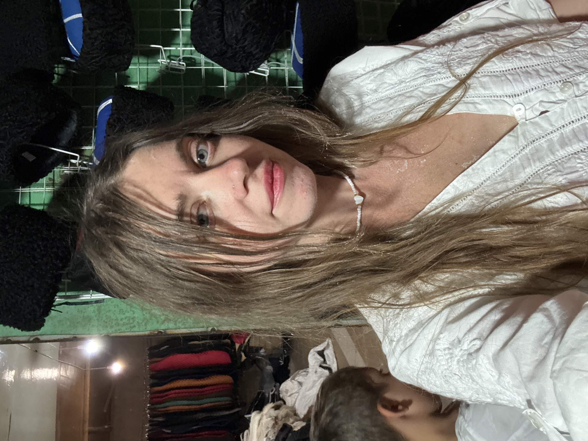 A woman with long, light brown hair is taking a selfie in what appears to be a market stall. She is wearing a white embroidered blouse and a white beaded necklace with a heart-shaped pendant. Her skin appears to have some powdery residue on it. In the background, there are shelves displaying dark, furry hats, and a rack of colorful garments. To the left, a young boy with short brown hair is visible, partially obscured. The environment suggests a storage or retail space, possibly in a city like Sol-Iletsk, Russia, known for its salt mines and associated local commerce.
