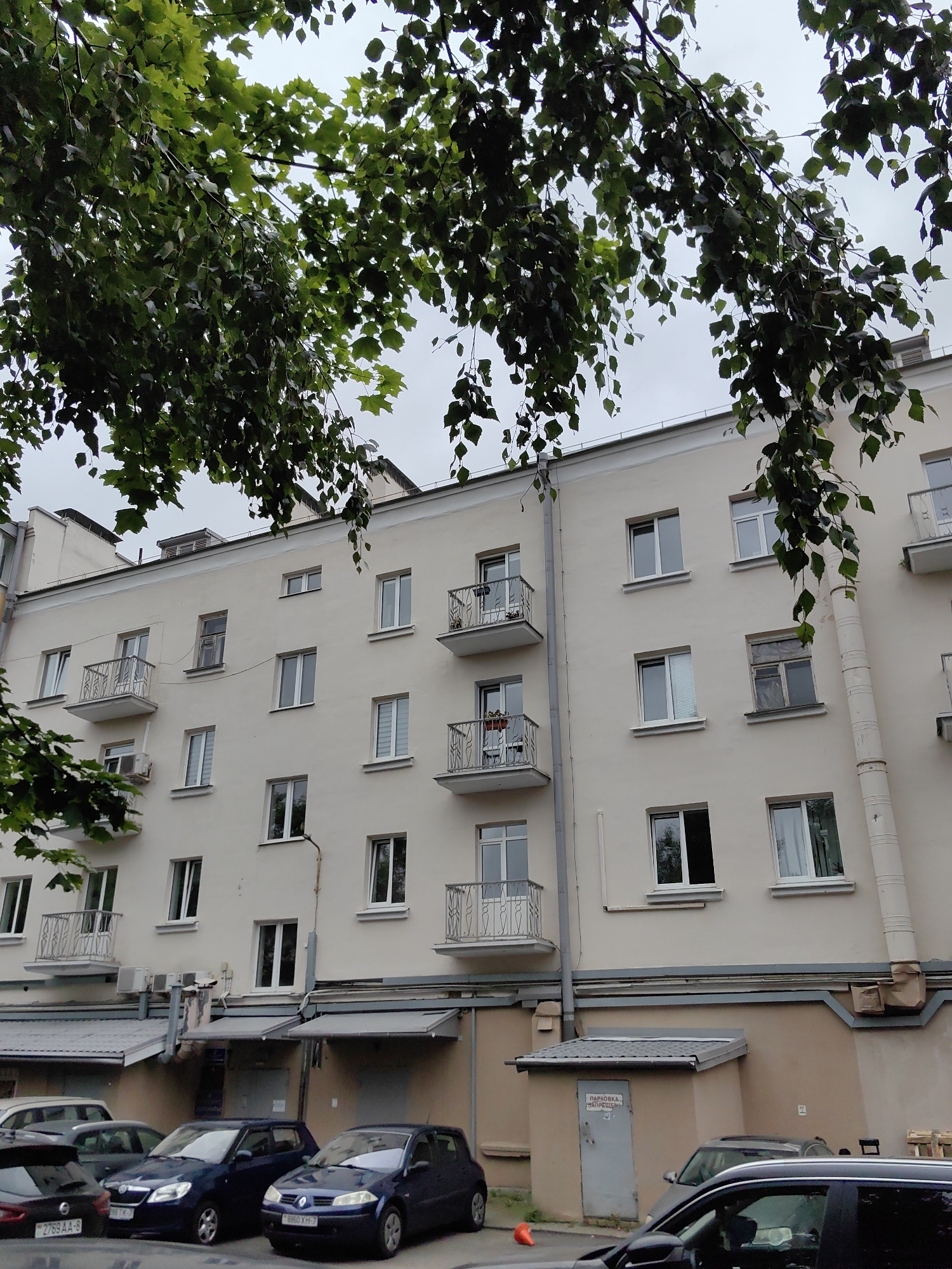 This is a ground-level, eye-level shot of the side of a beige apartment building in Minsk, Belarus. The building has at least five floors, with a series of windows and balconies on each level. Some windows are open, others are closed. There are several air conditioning units mounted on the building's exterior, and a large, cream-colored pipe runs vertically down the side of the building.
In front of the building, there are multiple cars parked, including a dark blue hatchback with a license plate that appears to read "8860 XH-7," a dark blue hatchback with a license plate "98 TK-7," and a dark gray SUV with a license plate "2759 AA-8." There is also a small, orange traffic cone in the foreground. The building is partially obscured by lush green foliage hanging from trees in the foreground, suggesting a warm season. The sky is overcast and gray.
