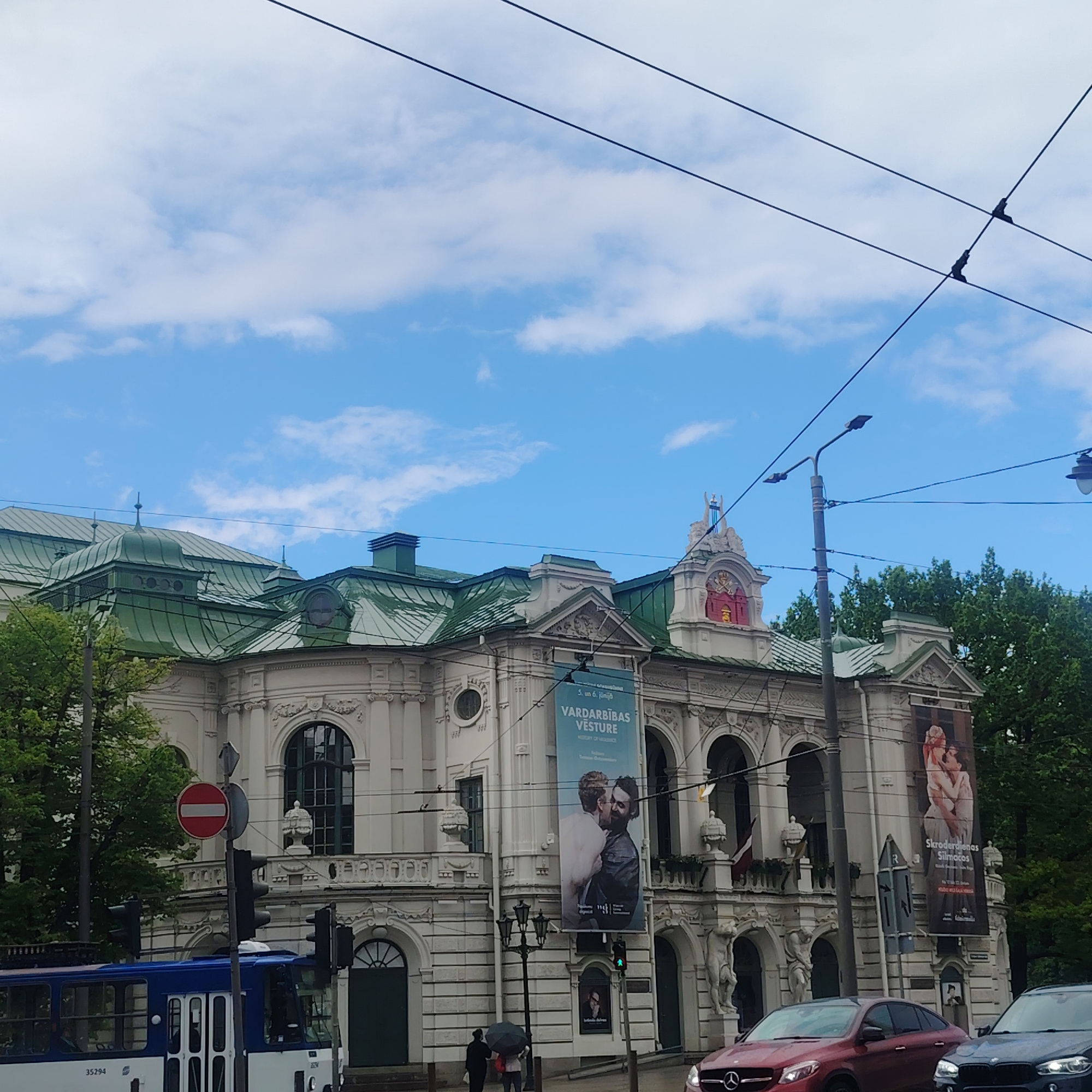The image shows the Latvian National Opera building in Riga, Latvia, on a day with a blue sky and scattered clouds. The building is white with a green roof. Banners advertising productions are displayed on the facade. A tram, cars, pedestrians, a traffic light, and overhead wires are visible in the foreground. The area appears to be a street with trees in the background.