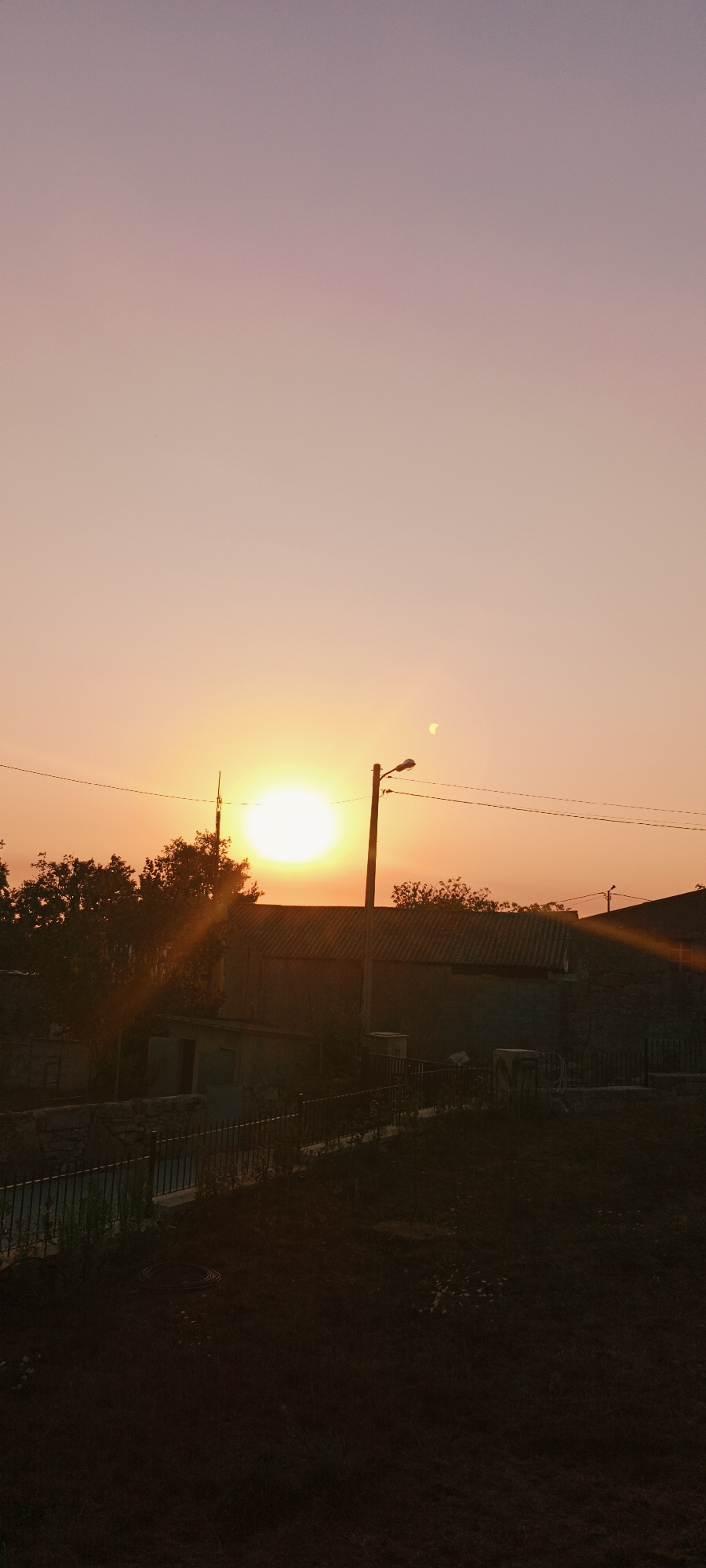 The image shows a sunset in the city of Adai, Spain. The sun is low in the sky, casting a warm glow over the scene. Silhouetted against the bright sky are trees, buildings, and utility poles. The sky is a gradient of orange and purple, transitioning to a lighter pink hue at the top. In the foreground, the ground is dark and appears to be covered in grass and some debris. A black metal fence runs horizontally across the mid-ground, separating the foreground from a paved area or a yard beyond. To the left of the image, there is a low-lying building with a dark roof and a small structure in front of it with an open doorway. In the center of the image, there is a larger building with a corrugated roof. A street light stands tall on a utility pole to the right of center, and overhead power lines crisscross the sky. A sliver of the moon is visible in the sky to the right of the sun. The overall impression is a peaceful evening scene in a semi-urban or rural setting within Adai.