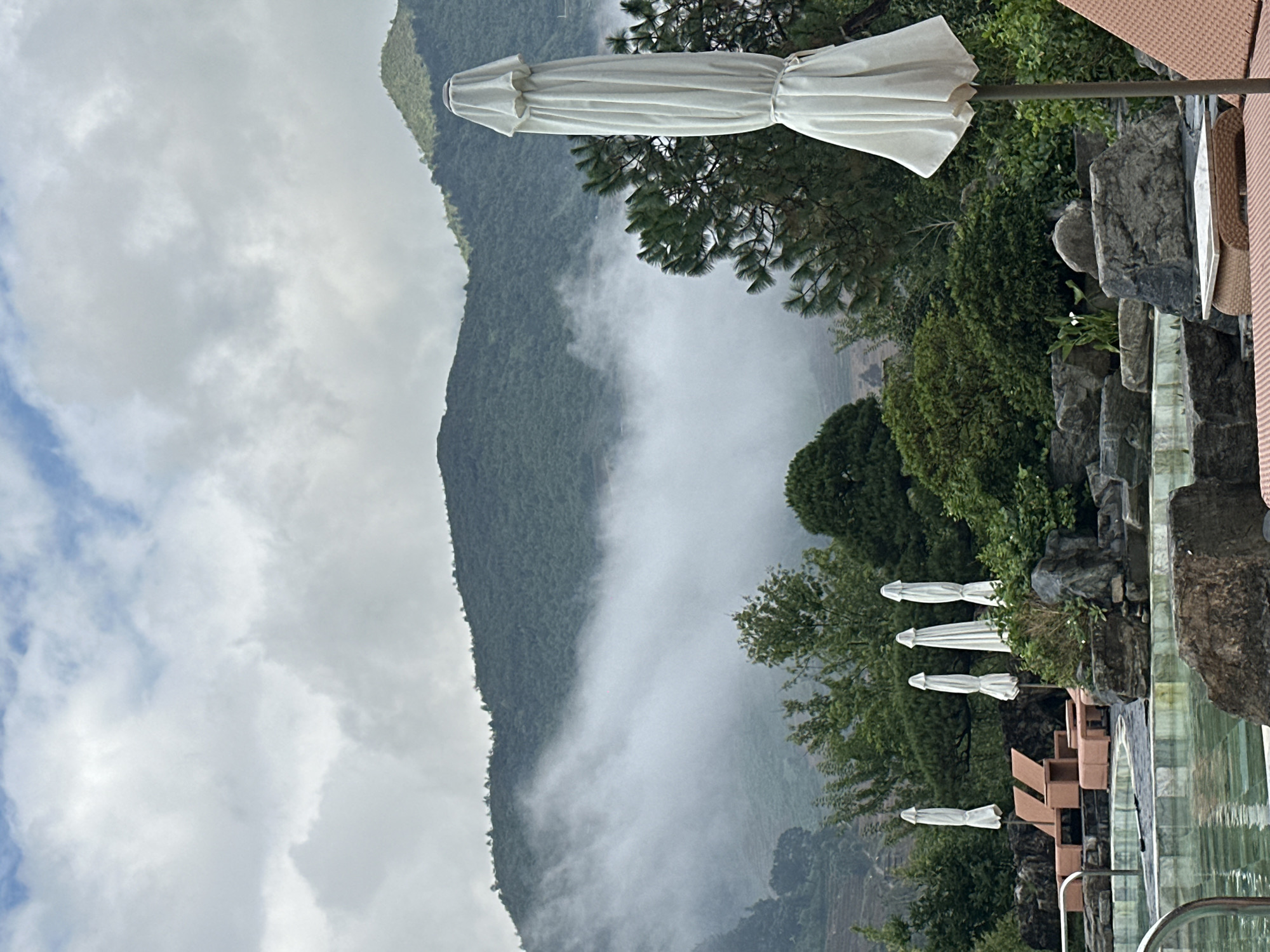 This image depicts a mountainous landscape and a resort area in Púng Luông, Vietnam, under an overcast sky. The upper portion of the frame shows a sky filled with diffuse white and light grey clouds. Below the sky, several large, green, heavily forested mountains dominate the background. A prominent band of white mist or low-lying clouds partially obscures the mid-slopes of the mountains, extending horizontally across the central part of the image. On the far left, hints of terraced topography are visible on a mountain face.
In the midground and foreground, a landscaped area features a swimming pool with light green water on the lower left. Surrounding the pool and extending towards the right are numerous large, grey rocks and diverse green vegetation, including coniferous and deciduous trees, and various bushes. Several white, folded outdoor umbrellas are scattered throughout this area. Specifically, two unoccupied orange-brown woven lounge chairs with accompanying footrests are positioned to the left of the pool. To their right, three more white, folded umbrellas stand upright. In the immediate foreground, a large, white, folded outdoor umbrella occupies the right side of the frame, partially obscuring the vegetation behind it. The top right corner of an orange-brown lounge chair is visible at the bottom right edge of the image. No people are present.