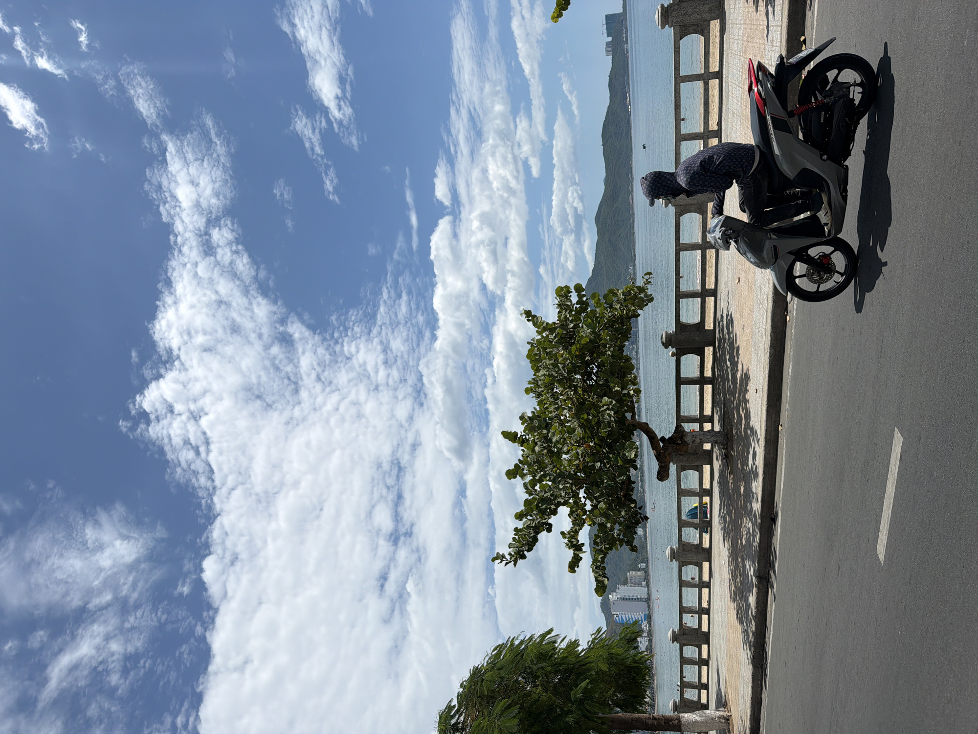 An overcast, bright day is captured from a street-level perspective looking towards the ocean in Nha Trang, Vietnam. A person wearing a dark hooded jacket and dark pants rides a grey scooter on the asphalt road. The scooter has red accents on the rear. The person is angled away from the camera, facing towards the sea.
Behind the scooter, a concrete promenade runs parallel to the ocean, bordered by a decorative stone railing. Trees, including a distinctive, gnarled tree with dark green foliage, cast shadows on the promenade. The ocean is a calm blue, with distant green mountains forming the backdrop. Scattered on the water are a few small boats, and a yellow and blue object, possibly an umbrella or beach shelter, is visible on the sandy beach behind the railing. Several tall buildings, characteristic of a coastal city, are visible against the mountainside. The sky is a bright blue, filled with fluffy white cumulus clouds. The overall scene depicts a typical day in a Vietnamese coastal city, with a focus on the activity on the road and the scenic view of the sea and mountains.