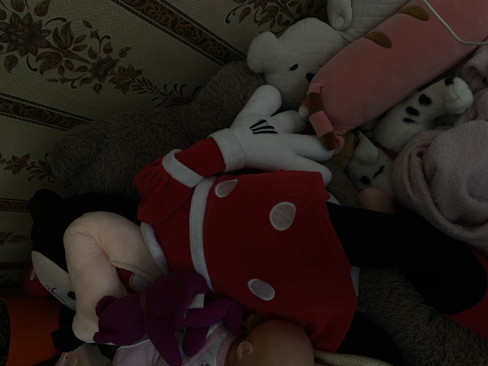 The image shows a cluttered collection of stuffed animals on a couch. A large Mickey Mouse plush is prominent in the center, wearing a red Santa-style outfit with white trim and white polka dots. One of Mickey's white, cartoon-style gloves is visible. To the left of Mickey, a small purple stuffed bear is partially obscured. Behind Mickey, a large, fuzzy brown teddy bear is visible. To the right of Mickey, a white quilted stuffed animal, possibly a bear or bunny, is visible. In the foreground, a long pink plush toy with brown spots lies horizontally. A stuffed animal with black spots, resembling a dog or cat, is partially underneath the pink toy and a pink blanket. The background features a floral patterned wallpaper. The scene is dimly lit, suggesting it is indoors, likely in a residential setting. There are no people visible in the image. The objects are piled on top of each other on a couch. This scene is not specifically identifiable as being in Lomonosov, Russia; it appears to be a typical indoor domestic scene.
