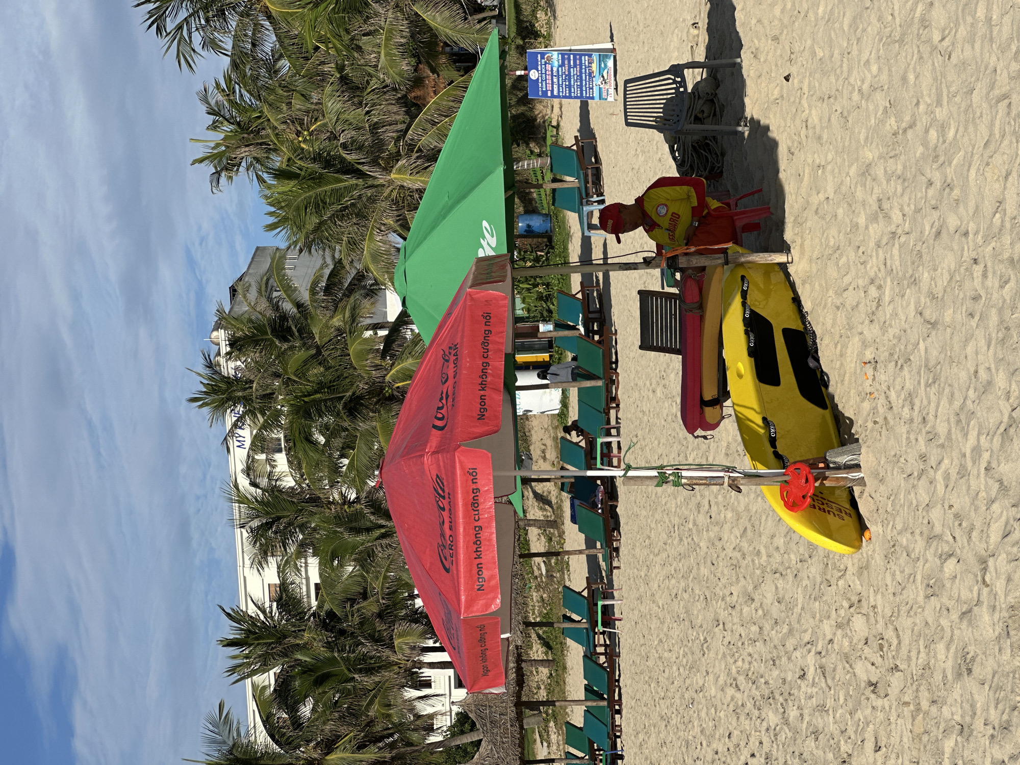The image depicts a beach scene in Đà Nẵng, Vietnam, under a clear blue sky with scattered clouds. In the foreground, two large beach umbrellas stand on the sand: one is red with a Coca-Cola Zero Sugar logo and Vietnamese text, and the other is green. Beneath the umbrellas, a lifeguard wearing a yellow and red uniform sits on a red plastic chair, looking at their phone. Next to the lifeguard are a yellow rescue board and a red flotation device. Several lounge chairs are arranged on the sand, some occupied by unseen individuals. In the background, palm trees line the beach, with a multi-story white building visible behind them. A blue barrel and a sign with Vietnamese text are also present on the beach.