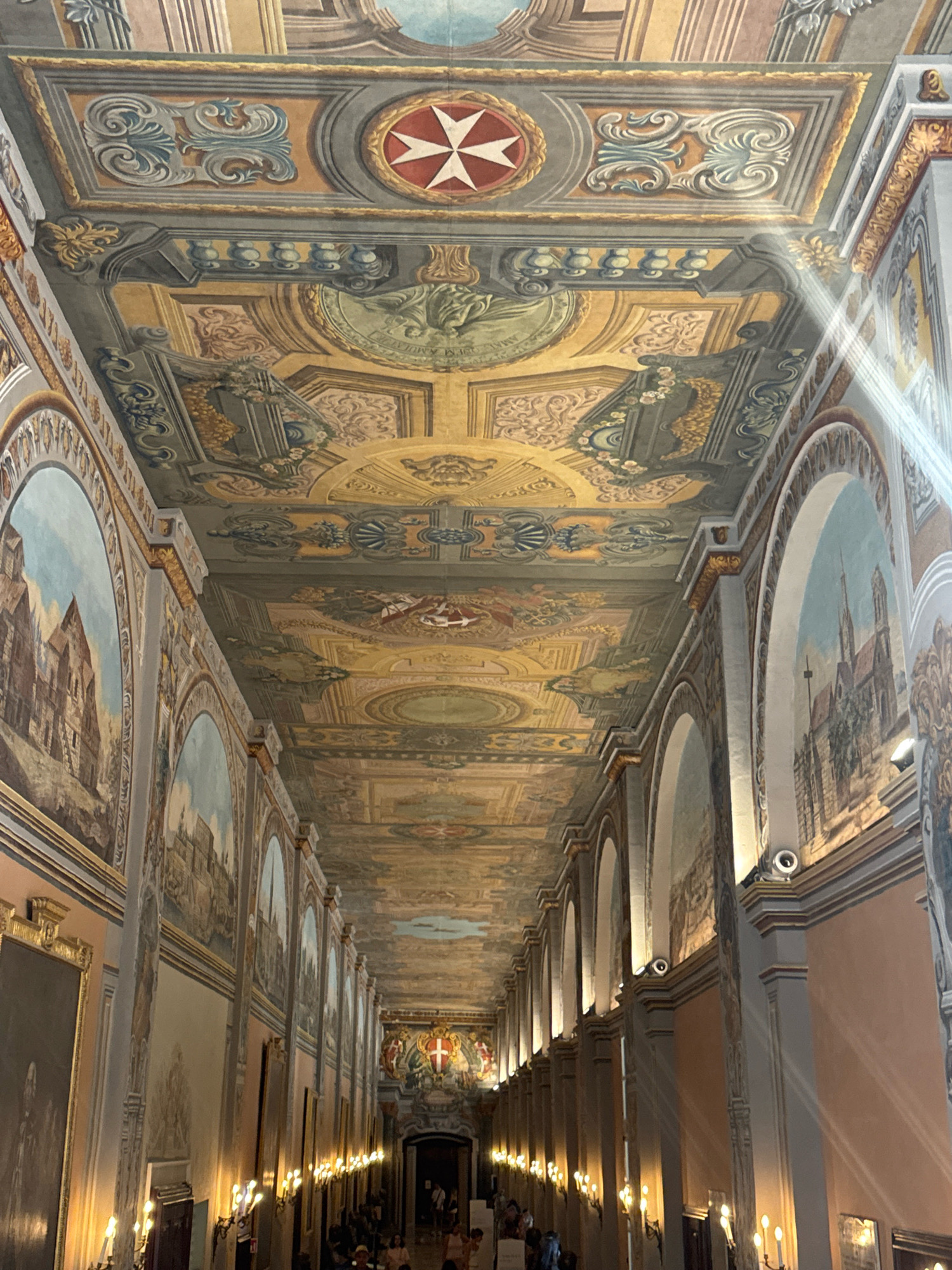 The image depicts the interior of a long corridor, likely a historical building, with a highly ornate ceiling painted with intricate frescoes and decorative moldings. The ceiling features a prominent Maltese cross in a circular emblem at its highest point, surrounded by a complex array of architectural motifs, floral patterns, and possibly mythological figures. The walls are adorned with large arched paintings, which appear to be landscapes or cityscapes. The corridor is illuminated by artificial lighting, with sconces lining the walls and ceiling lights, casting beams of light that are visible in the upper right portion of the image. Several people are visible at the far end of the corridor, suggesting this is a public space, possibly a museum or a historical site within Valletta, Malta. The overall impression is one of grandeur and historical significance.