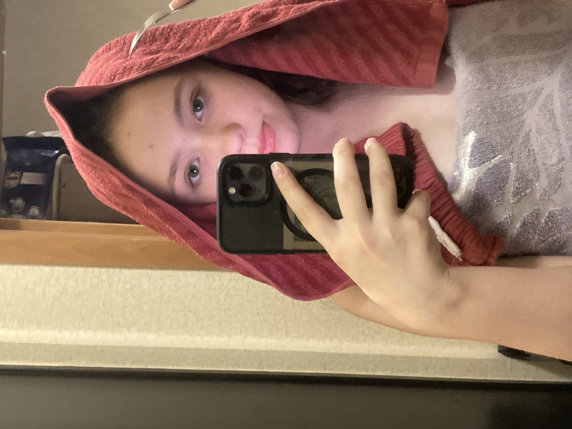 A girl is taking a selfie in a mirror, possibly in a bathroom setting. She is holding a black smartphone in her right hand, which is partially obscuring her face. She is wearing a red towel draped over her head and shoulders, creating a hood-like effect. A metallic object, possibly a hair clip or fork, is visible above the towel on her head. The reflection in the mirror shows a counter with items on it. The counter has a light-colored finish, with a dark-colored cabinet beneath it. Another towel with a white embroidered pattern lays on her chest.