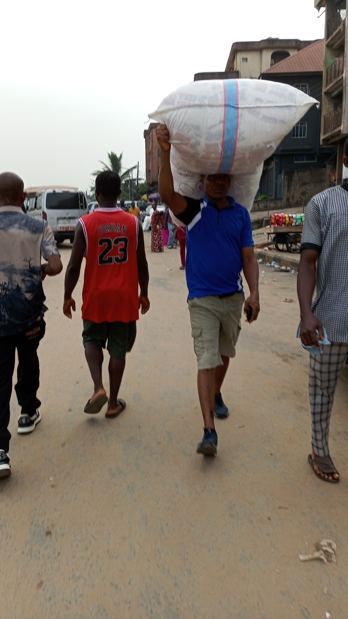 The photo, taken in Onitsha, Nigeria, depicts a street scene. A man carries a large white sack on his head. He wears a blue polo shirt and khaki shorts. Three other men walk on the road. One wears a black and white shirt and black pants; another wears a red "Jordan 23" jersey and green shorts; the third wears a patterned shirt and plaid pants. In the background, there's a white Toyota HiAce van, buildings, and other pedestrians. A cart laden with packaged drinks is visible on the right side of the street. The road surface is rough.