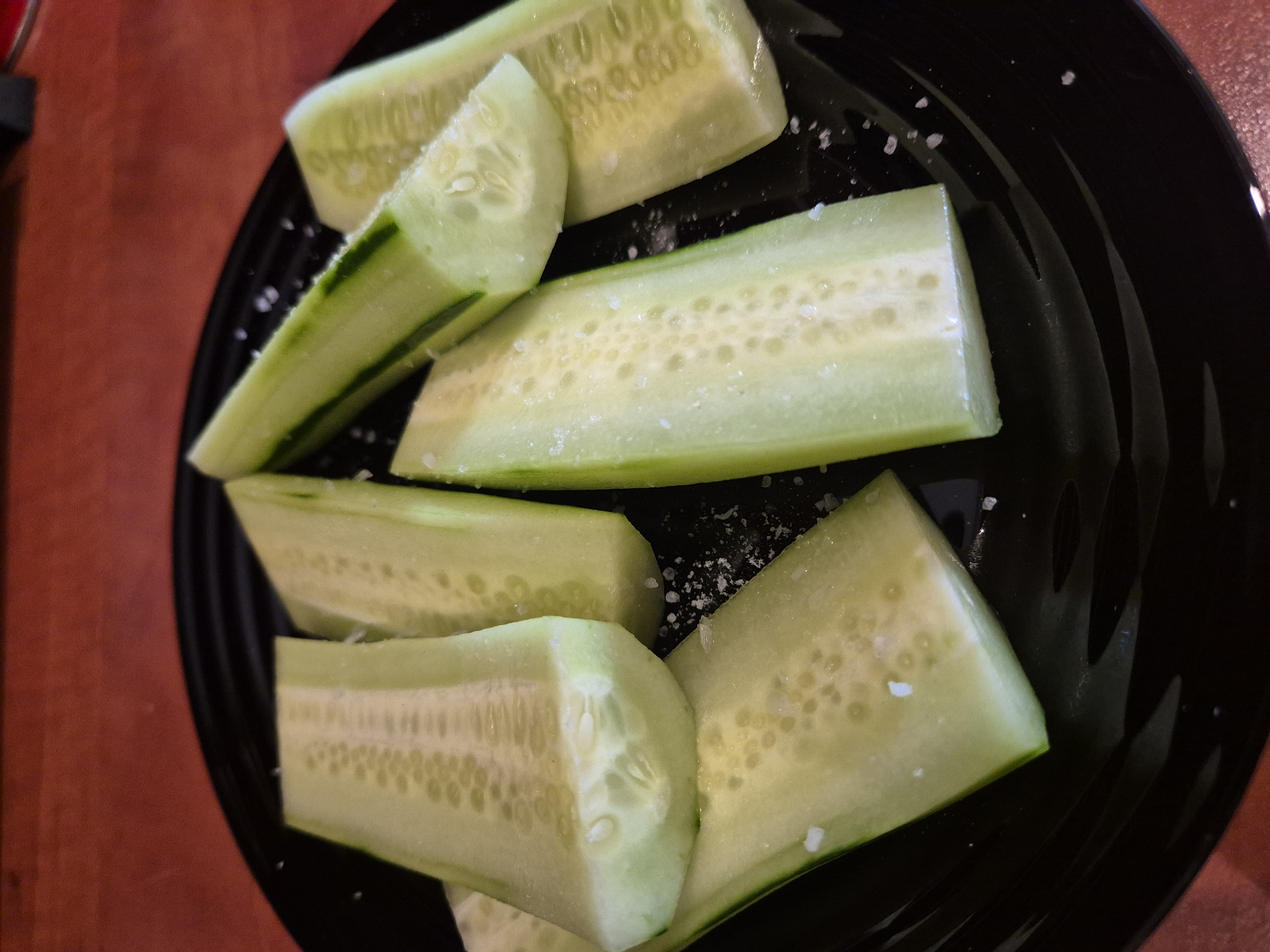 A close-up, overhead shot shows several slices of cucumber on a black plate. The cucumber slices are cut lengthwise, with some of the seeds visible in the center. They are sprinkled with salt. The plate is on a reddish-brown surface, possibly a wooden table. The lighting is somewhat dim, creating soft shadows.