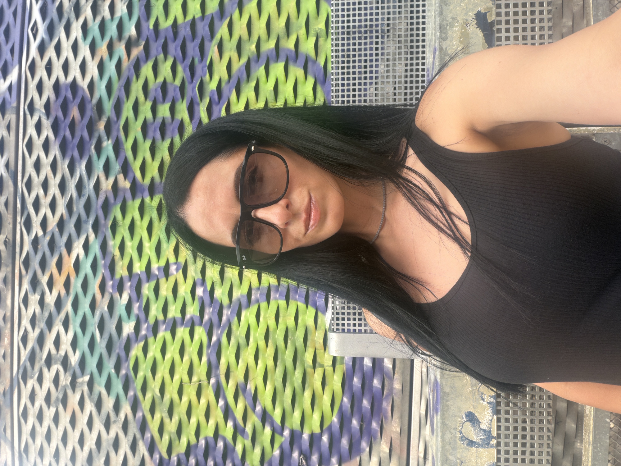 A woman with long, dark hair and sunglasses is captured in a selfie, leaning slightly to her right. She is wearing a black, ribbed tank top and a delicate necklace. Her gaze is directed towards the camera. She is seated on a metal bench with a checkered pattern, and behind her is a textured wall with abstract graffiti in shades of green and purple. The backdrop suggests an urban outdoor setting. The specific location is the city of Corsico, Italy.