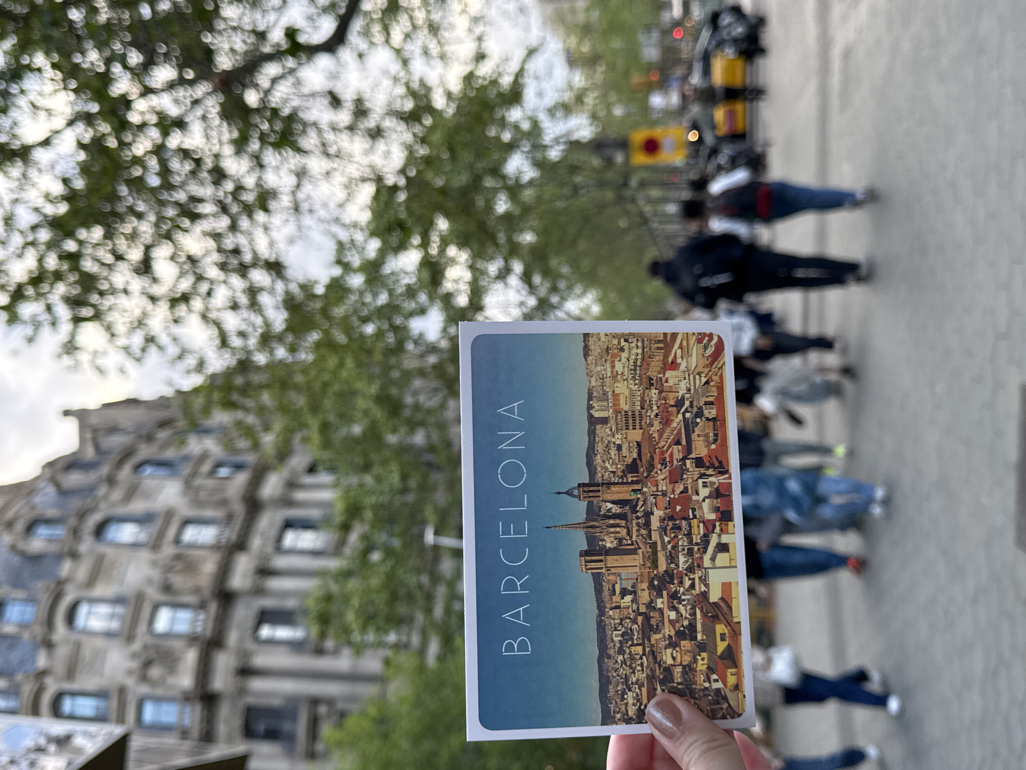 A postcard of Barcelona held by a hand is superimposed on a blurry street scene in Barcelona, Spain. The postcard depicts a panoramic view of the city, with the Sagrada Familia prominently visible. The background features a blurred street scene with trees, people walking on the sidewalk, a building with many windows, and what appears to be traffic lights and a yellow sign on the street. The overall image suggests a tourist holding up a postcard to compare it to the actual location.