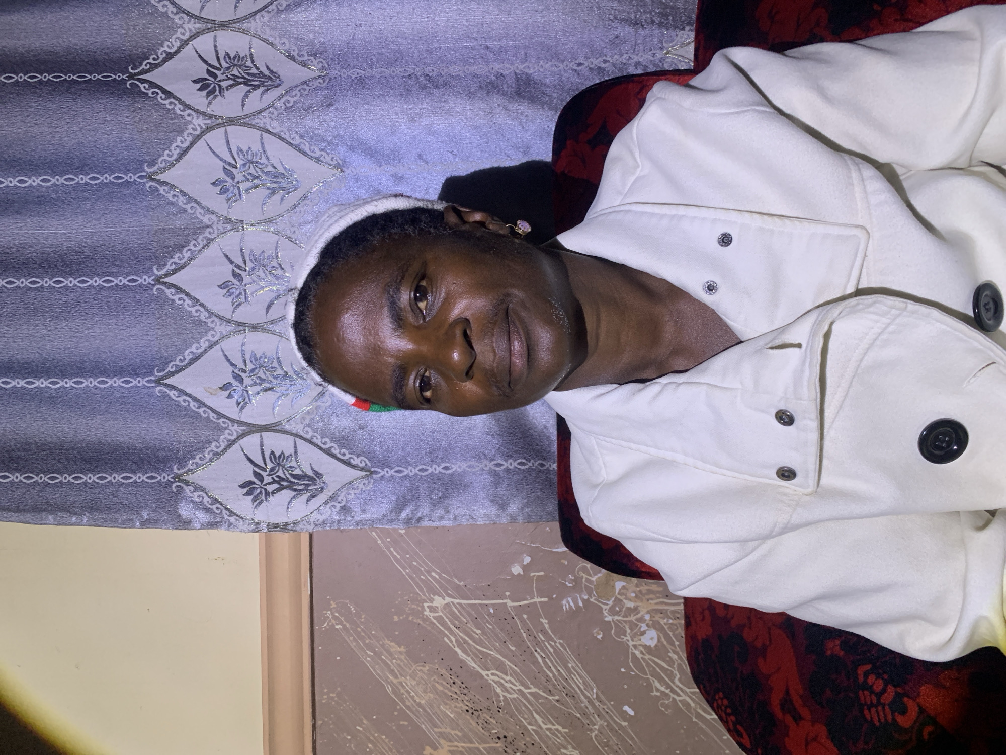 A portrait shows a Black woman with dark skin, short dark hair, and a light-colored head wrap, seated in a chair in Barakin Sabawa, Nigeria. She is wearing a white coat. The background consists of a patterned curtain with a floral design. The room appears to be dimly lit, with a spotlight on the left side of the frame. The woman is looking directly at the camera with a neutral expression. The overall impression is one of a simple, intimate setting.