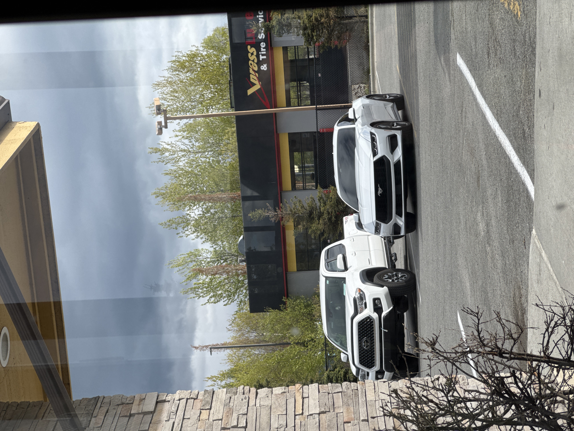 The image shows a view from inside a building, looking out at a Vopass Tire & Tire Service store. Parked in front of the store are two white vehicles: a Toyota Tacoma pickup truck and a Ford Mustang sports car. The sky is overcast. Based on the presence of a Vopass Tire & Tire Service store, the location is likely in Anchorage, Alaska, United States of America.