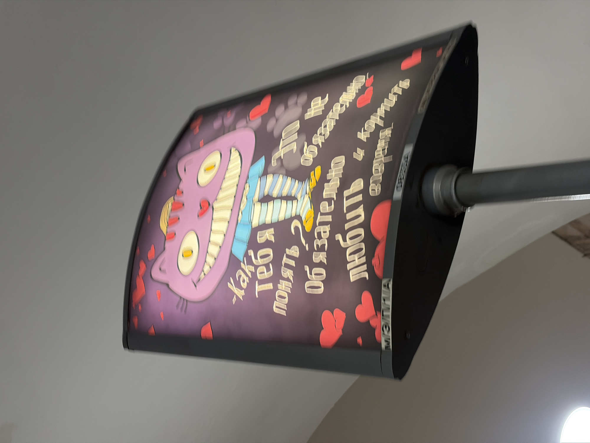 The image displays an illuminated, curved sign featuring a vibrant purple Cheshire Cat with a wide, toothy grin and striped legs, characteristic of the character from "Alice's Adventures in Wonderland." The background of the sign is a dark purple, dotted with numerous red heart shapes of varying sizes.
The main text on the sign is written vertically in Cyrillic, in a stylized golden font. It reads: "Как тебя понять? Обязательно любить И кормить." This translates to "How to understand you? Must love And feed," a popular quote associated with the Cheshire Cat. On the right-hand edge of the sign's black frame, the word "МЕЛГИА" (MELGIA) is visible in white.
The sign is mounted to a light grey ceiling by a grey metal pole extending from its right side, suggesting it is a decorative element in an indoor public space. The lighting appears to be artificial, coming from within the sign itself and illuminating the artwork and text. The surrounding environment consists of plain, light-colored walls and ceiling, providing a neutral backdrop that highlights the colorful sign. The overall aesthetic is whimsical and romantic, likely indicating a themed establishment like a cafe, bookstore, or entertainment venue in Saint Petersburg, Russia.