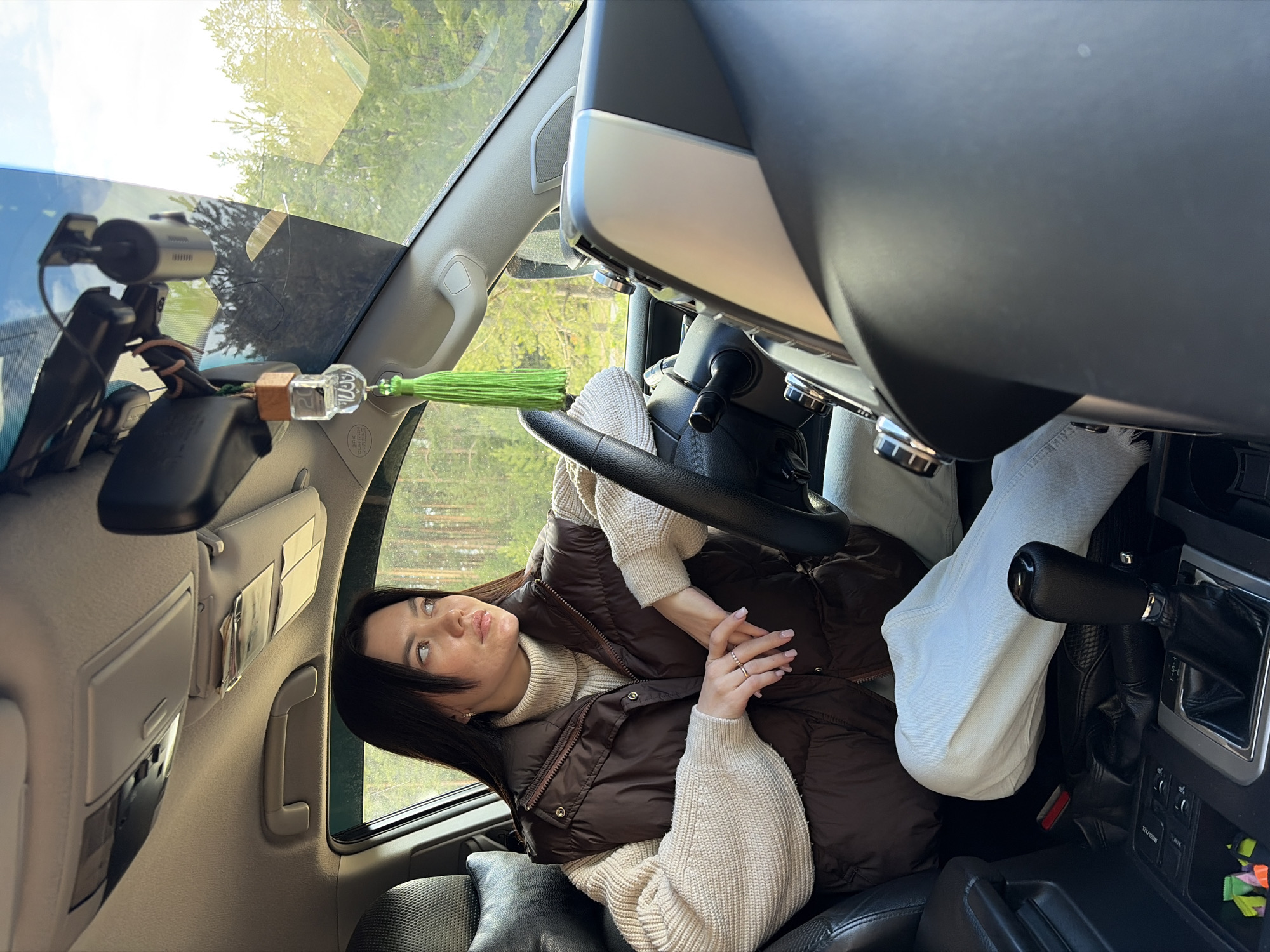 A young woman with long dark hair is seated in the passenger seat of a car, looking upwards and slightly to her right with a pensive expression. She is wearing a cream-colored turtleneck sweater and a brown puffer vest, with light-colored pants. Rings are visible on her right hand, which is clasped in front of her.
The interior of the car is visible, featuring a black steering wheel, a dashboard with silver accents, and black leather-trimmed seats. A dashcam is mounted on the windshield, near a visible crack or scratch. Hanging from the rearview mirror area is a car charm or air freshener with a wooden base, a clear faceted cube displaying the number "2", and a vibrant green tassel. The sun visor on the passenger side is flipped up, holding several cards. A partially visible "SRS AIRBAG" label can be seen on the pillar. Below the gear shifter, which has a black leather boot, there are charging ports labeled "12V/220W". Colorful candies or small items are scattered in a compartment nearby.
Outside the car, through the windshield and side window, lush green trees and a bright, clear sky are visible, suggesting a sunny day in a natural or semi-natural environment. The scene takes place in Chaikovsky, Russia.