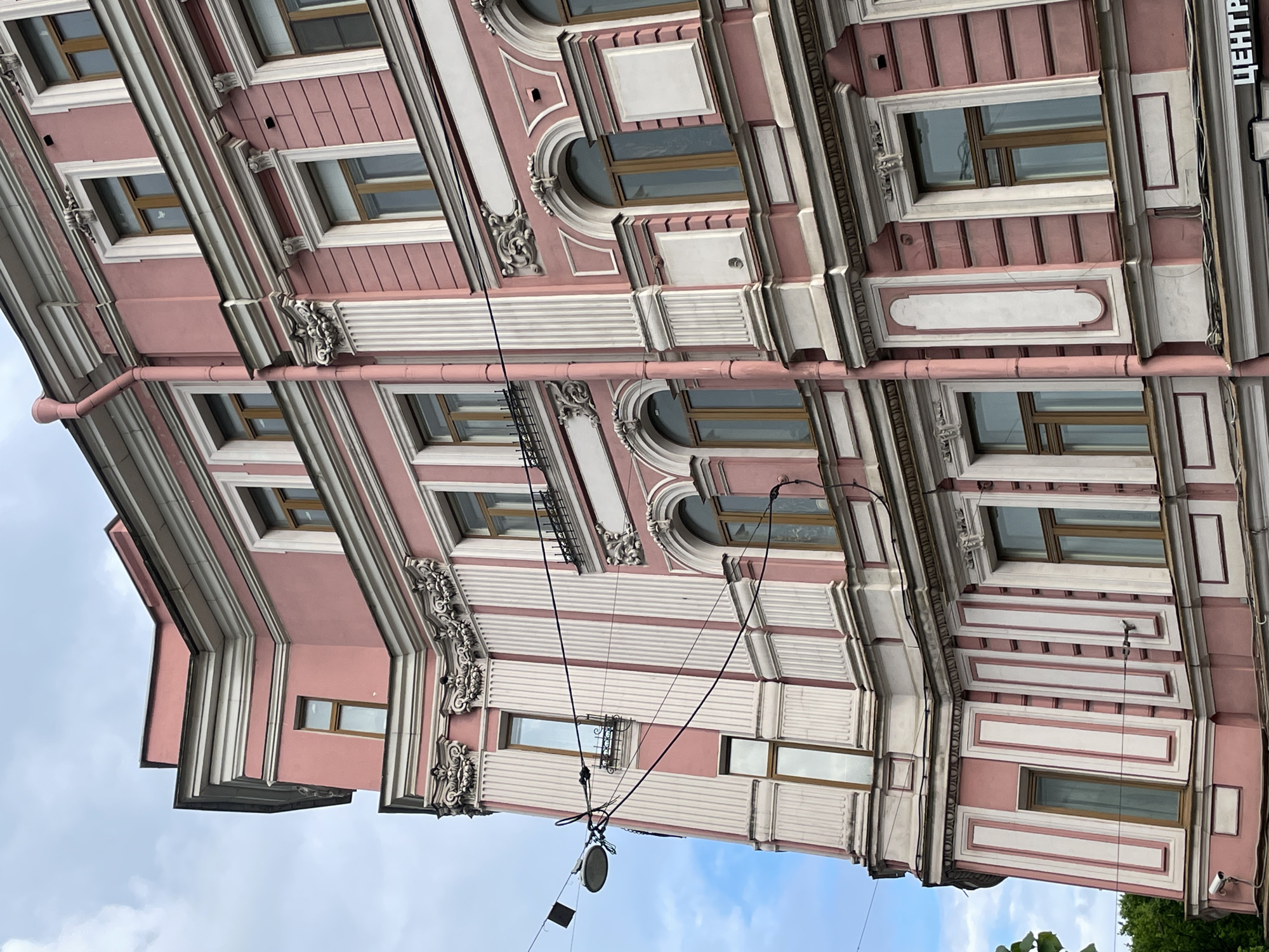 The image, presented rotated 90 degrees clockwise, showcases the ornate facade of a multi-story building, characteristic of grand European architecture, located in Saint Petersburg, Russia. The building is predominantly painted in a salmon or light pink hue, beautifully contrasted with extensive off-white and light gray architectural detailing.
The facade is richly adorned with classical elements, suggesting a Neoclassical or Baroque revival style. Vertical pilasters, fluted columns, cornices, and intricate moldings frame the windows and define the building's structure. Windows are a mix of rectangular and elegantly arched designs, often featuring brown frames. Decorative motifs, including floral and scrollwork, are prominent below window sills and at the capitals of pilasters. Sections of the wall exhibit textured horizontal grooves, adding visual interest.
A salmon-colored drainpipe runs vertically down the left side of the building. Multiple black electrical wires crisscross in front of the facade, from which a circular street lamp fixture and a small grid-like antenna are suspended. A small security camera is visible on the lower right portion of the building.
The background shows an overcast sky with scattered clouds, indicating daytime. Green foliage from a tree can be seen in the bottom right corner. On the far right section of the building, a vertical sign clearly displays the Russian word "ЦЕНТР" (TSENTR), meaning "CENTER." The building appears well-maintained, though some minor weathering is visible. No people or discernible activities are present; the focus is entirely on the architectural details.