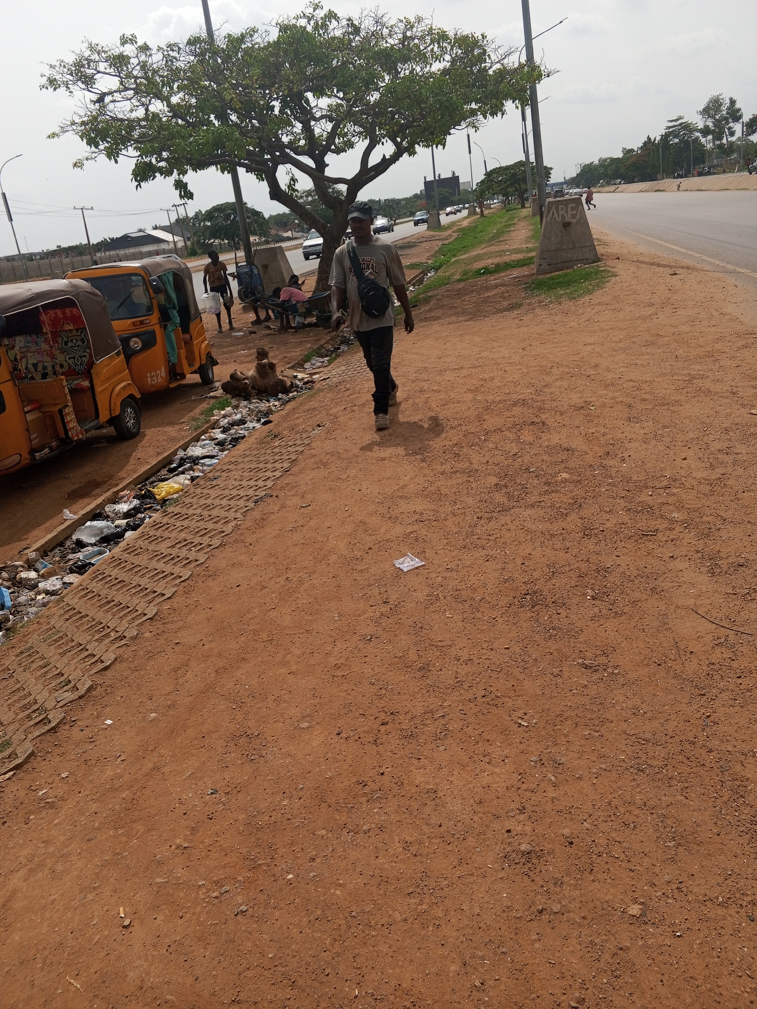 The image depicts a street scene in Abuja, Nigeria. The ground is reddish-brown dirt. A drainage ditch filled with trash runs along the edge of the dirt path. Two yellow auto rickshaws are parked along the ditch. Several people are standing around. One man is walking away from the others. A tree provides shade in the background. Cars and buildings can be seen in the distance.
