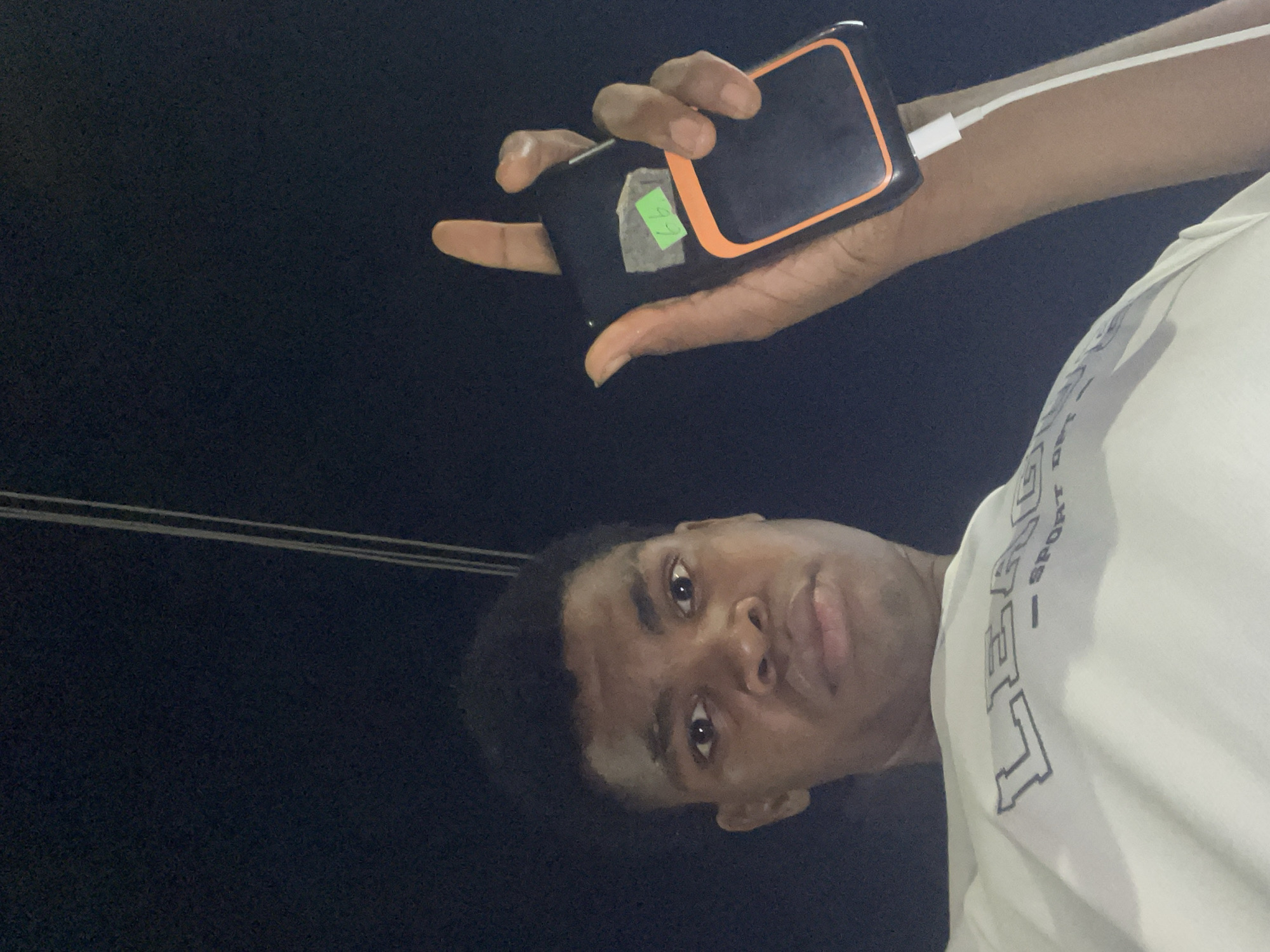 A person is holding a black smartphone with an orange border and a white charging cable. The phone has a green sticker on its back. The person is wearing a white t-shirt and is located outdoors at night in what appears to be Ado, Nigeria. The presence of power lines further confirms an urban location.