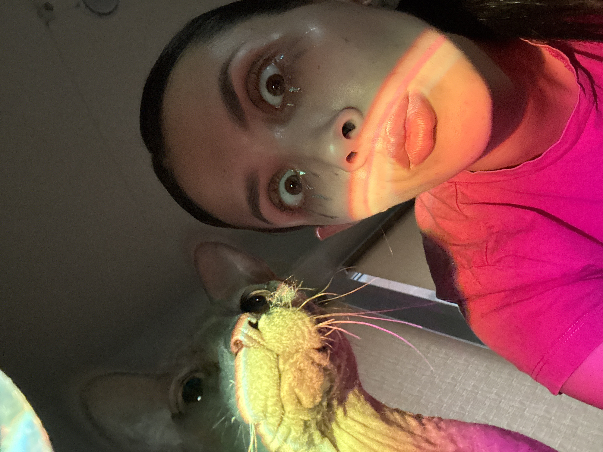 A low-angle selfie features a woman with dark hair pulled back, wearing a bright pink shirt, alongside a hairless cat. The woman has light skin, brown eyes, and her face is illuminated with a reddish-orange light. The cat is looking up towards the camera with a wrinkled face and visible whiskers. The background appears to be a room interior, with the ceiling visible in the upper left corner. The location in Votkinsk, Russia, cannot be determined from the provided image.