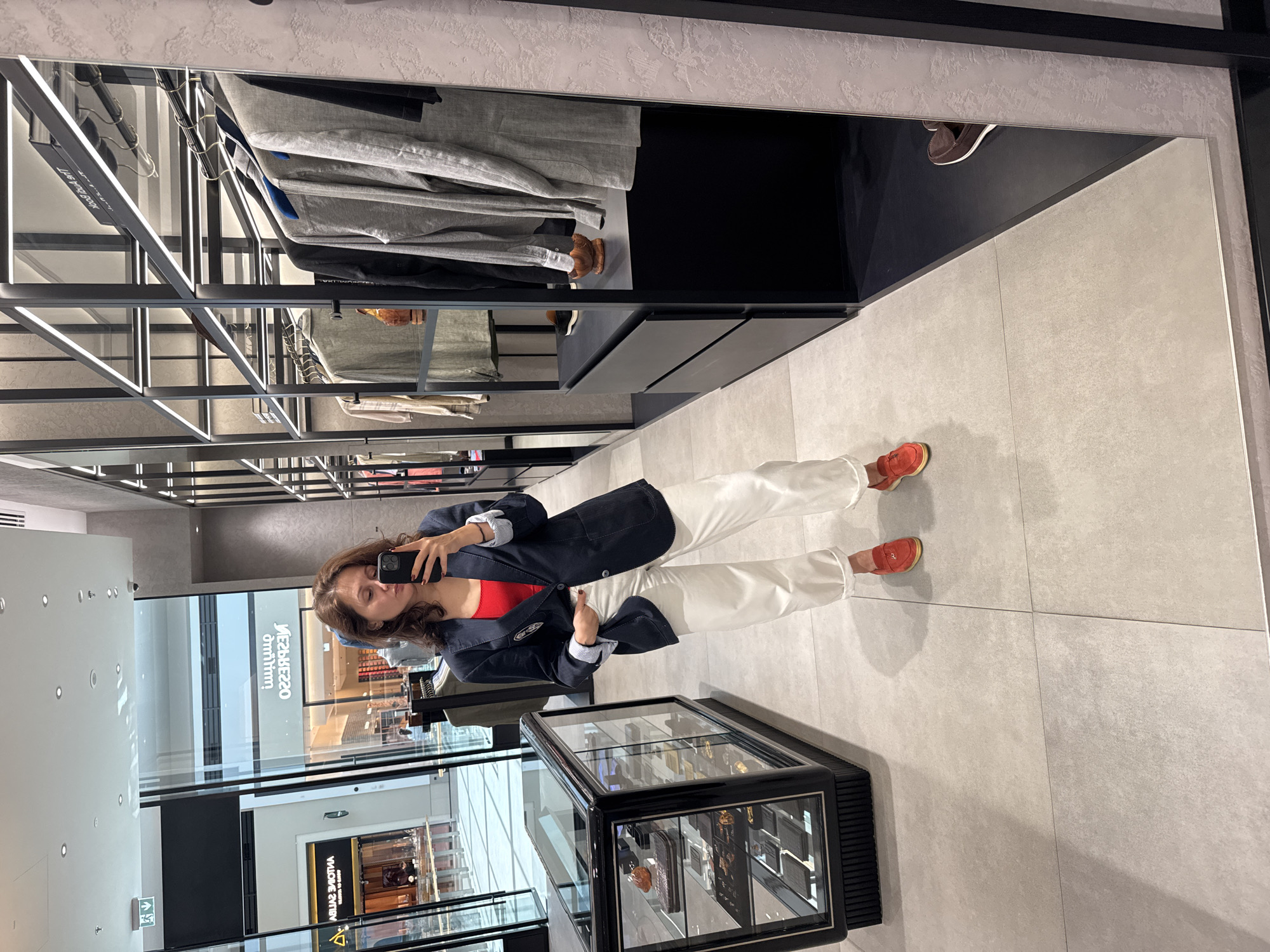 A woman is taking a selfie in a retail store. The store is located in Dubai, United Arab Emirates, as "ARMANI CAFFE" is visible. The woman is wearing a red top, white pants, and a navy jacket. She is standing in front of a display case. The store has clothing racks in the background. The floor is made of large gray tiles.