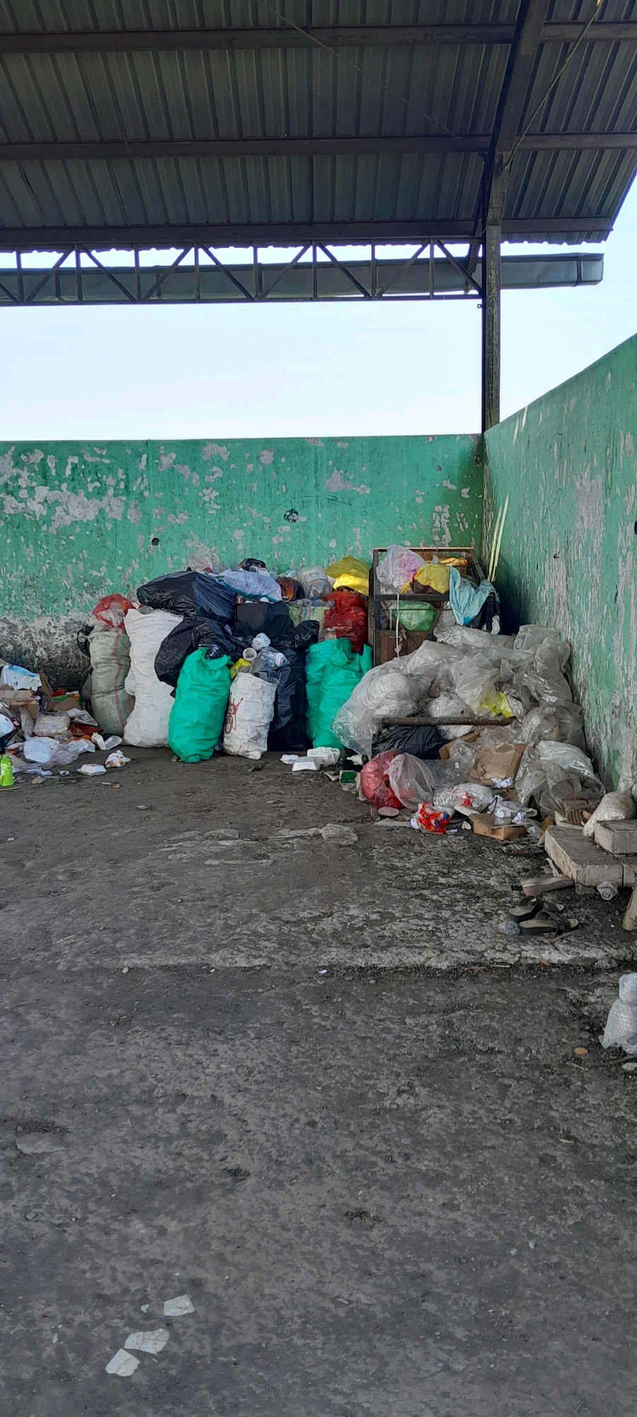 The image depicts an outdoor area under a metal roof structure, likely a recycling or waste collection point in Bendungan, Indonesia. A large pile of assorted garbage bags, predominantly black and white, along with several bright green and red bags, is consolidated against a peeling, light green concrete wall. Loose trash, including plastic bottles, paper scraps, and cardboard, is scattered on the ground. To the right of the main trash pile, a metal cage-like structure holds some colorful plastic bags, and more plastic-wrapped waste is piled high. The ground is concrete and appears weathered and dirty. In the background, a lighter colored wall is visible, and the sky is a pale blue. No people are present in the scene.