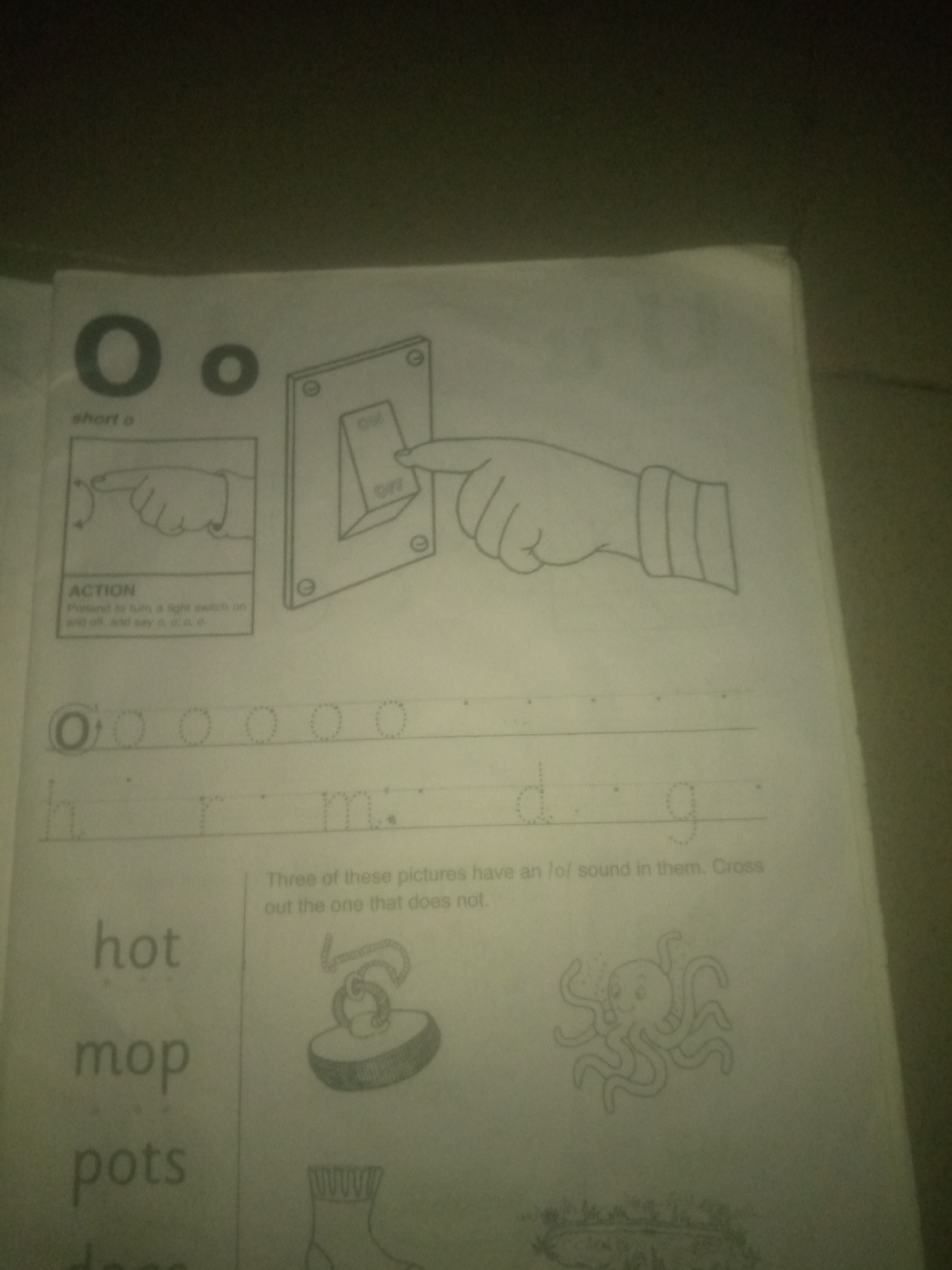 The image displays a page from a children's learning worksheet. At the top, there are large and small letter "O"s, with the text "short o" below. To the right, an illustration depicts a hand turning a light switch on and off, with the "ACTION" section below instructing the user to "Pretend to turn a light switch on and off, and say o, o, o." Below this, dotted lines show the letters "O", "h", "r", "m", "d", and "g", suggesting practice tracing these letters. Further down, on the left side, are the words "hot", "mop", and "pots". On the right side, there are three illustrations: a plunger or something similar, an octopus, and a sock. A caption above these illustrations reads, "Three of these pictures have an /o/ sound in them. Cross out the one that does not." The image quality is poor, with low resolution and poor lighting, making some details blurry. The scene is indoors, on a paper surface. There is no indication of the city of Jauro Yinu, Nigeria within the image content.