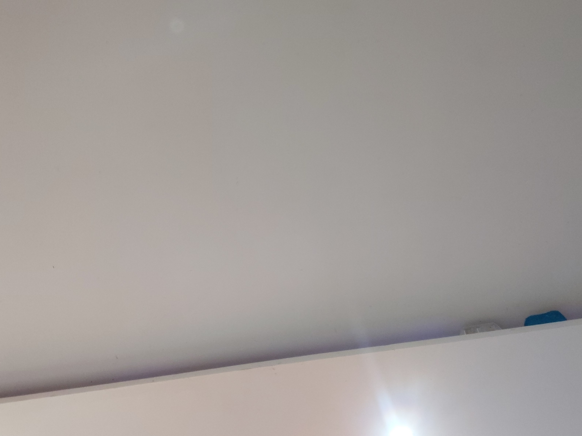 The media file displays an interior scene, primarily focusing on a light-colored, likely off-white or pale gray, ceiling or upper wall. The majority of the image is taken up by this smooth, unadorned surface.
Across the bottom third of the frame, a distinct horizontal line separates the upper surface from a lower, brighter surface. This lower area, possibly another wall or a reflective surface, is heavily illuminated by a strong light source, creating a prominent lens flare effect in the bottom right corner. This light source casts a bright glow that extends onto the edge of the upper surface.
Positioned on the edge of the upper surface, near the right side of the frame, are two small, inanimate objects. The first object, closer to the center, is a small, light-colored, possibly white or off-white rectangular item, which could be a small vent or a piece of plastic. Adjacent to it, further to the right, is a small, irregularly shaped blue object, possibly a sponge or a piece of fabric.
There are no people or discernible activities taking place. No visible text is present. The strong artificial light suggests an indoor setting. The image offers no specific visual cues to confirm the location in Santa Ana, Venezuela, but the context provided situates it there.