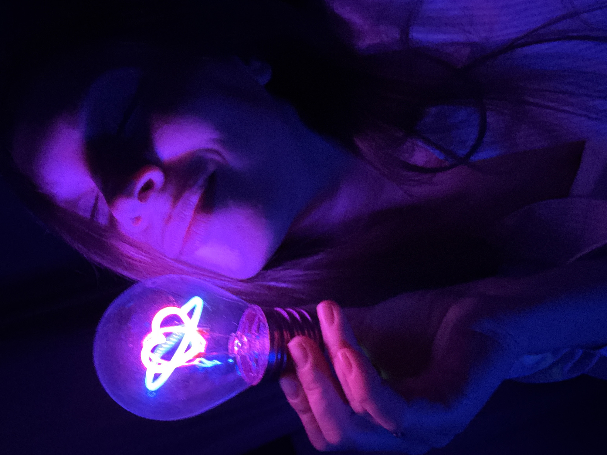 A person with fair skin and light hair is holding a lightbulb containing a neon art piece. The person is partially illuminated by the lightbulb, which emits a blue and pink glow. The individual is lying down, possibly in a dark room, with only parts of their face and upper body visible. The neon art inside the lightbulb is a design of intertwined shapes. No specific location in Budva, Montenegro, is discernable.