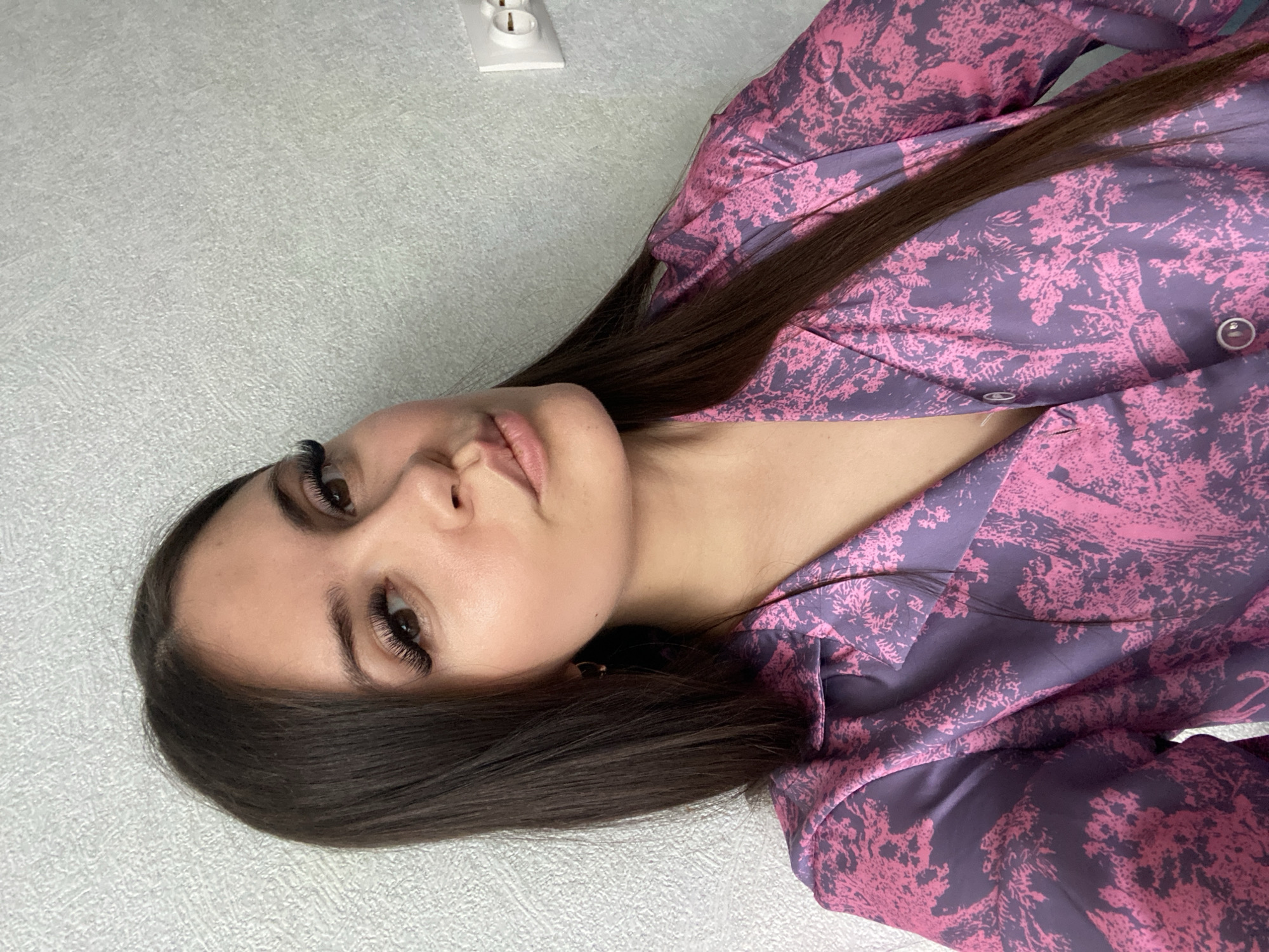 A woman with long, dark brown hair is centered in the frame, wearing a patterned pink and purple shirt. She has long eyelashes and is looking directly at the camera with a neutral expression. The background consists of a textured light gray wall with a white electrical outlet visible in the upper left corner.