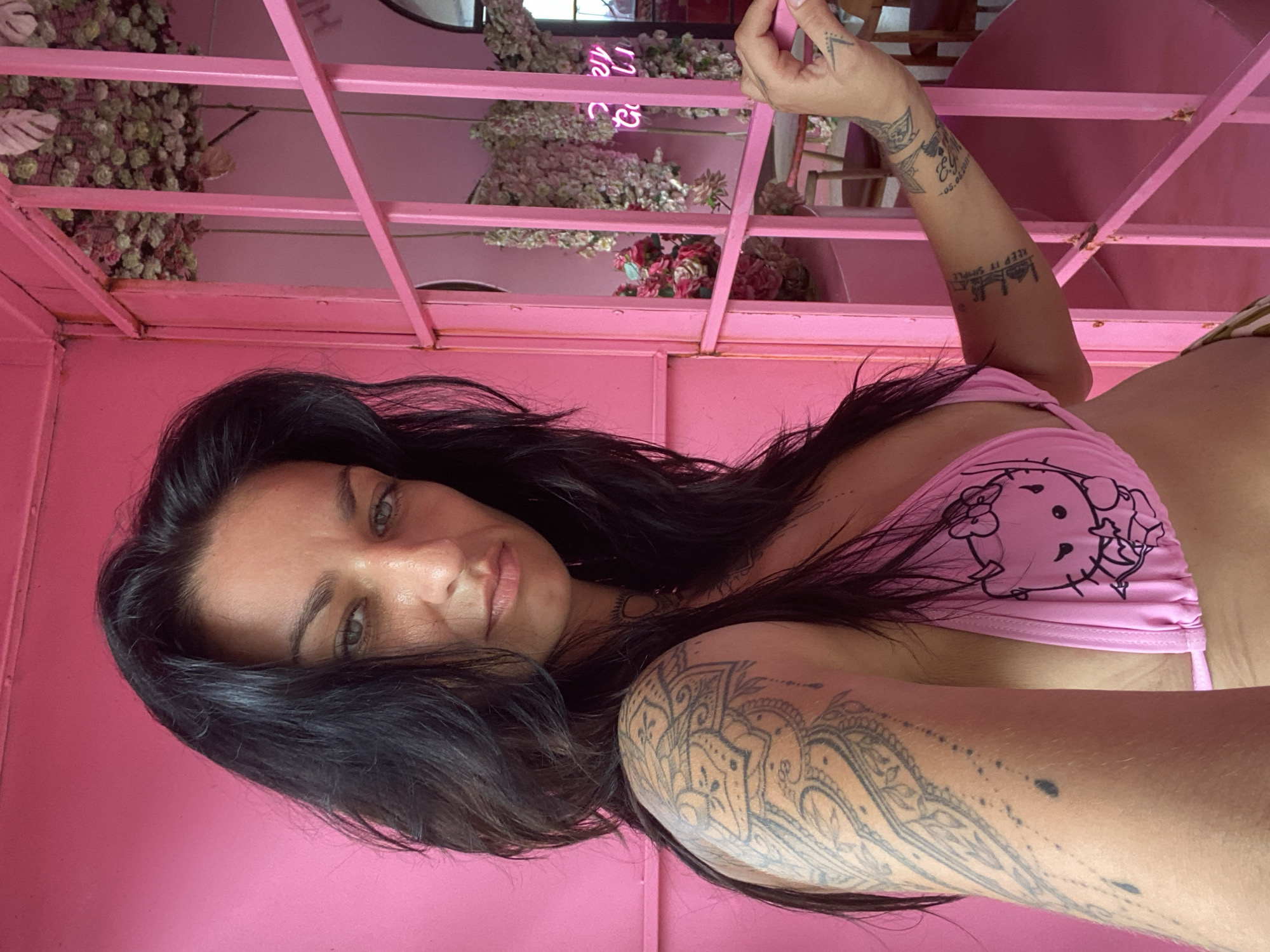 A woman with long, dark hair and light-colored eyes is captured in a portrait, looking directly at the camera with a calm expression. She wears a pink bikini top adorned with a black outline of a Hello Kitty design. Her arms are extensively tattooed; notable designs include a large, intricate mandala-like pattern on her left shoulder and upper arm, and the phrase "KEEP IT SIMPLE" on her left forearm. A partial date "-05.06.XXXX" and other smaller symbols are also visible on her upper arm, with a small arrow tattoo on her left hand.
The setting is vividly pink, with the woman positioned against a backdrop of pink walls and a structure made of pink metal bars, possibly a decorative canopy or cage. Hanging from this structure are numerous bundles of dried or artificial flowers and plants, primarily in light green and white tones, with some accents of pink and red. In the blurred background, a pink neon sign is partially visible, showing fragments of text like "ne" and "Co." The lighting appears bright and diffuse, indicating it is likely daytime. The overall scene suggests a vibrant, themed indoor or semi-enclosed environment, possibly a cafe or recreational spot in Tien Thanh Ward, Vietnam.