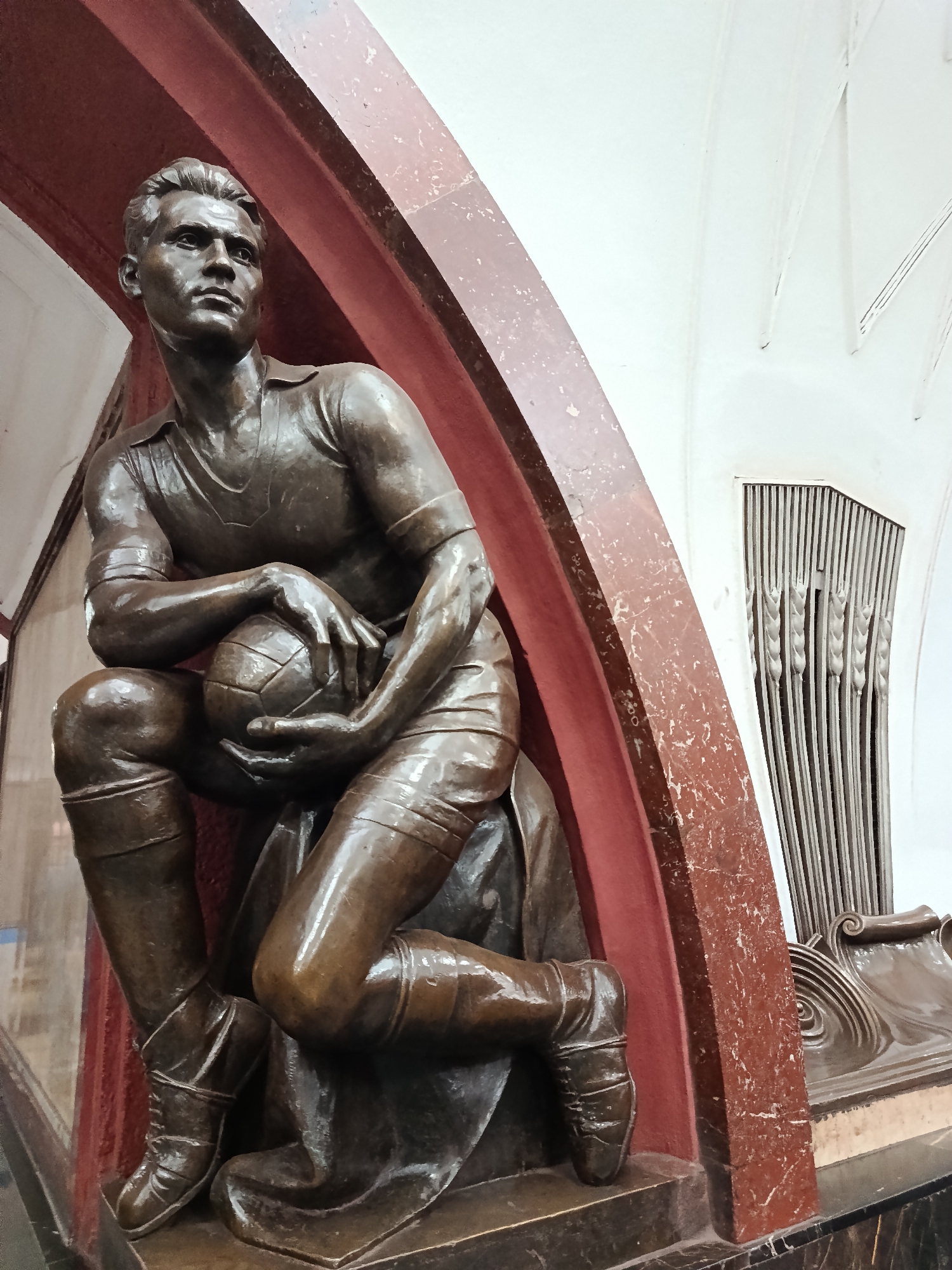 A bronze statue of a male athlete, likely a soccer player, is depicted in a kneeling or crouching pose, captured from a slightly low angle. The figure, cast in a polished, reddish-brown metallic material, possibly bronze, holds a classic panel-designed football in both hands, resting it on his bent left knee. He wears a short-sleeved collared jersey, shorts, and tall, laced boots. His face is turned upwards and slightly to his left, with a focused, determined, or contemplative expression.
The statue is set within a robust, reddish-brown archway or niche made of what appears to be veined marble or polished stone, displaying some scuffs and lighter specks. To the right of the archway, a light-colored (white or off-white) wall is visible, adorned with tall, vertical architectural details resembling stylized wheat stalks or reeds. Below these, a decorative relief with swirling patterns is present. The base supporting the statue and the archway is a dark, smooth material, possibly marble. The overall scene suggests an indoor setting, possibly a public space or monument hall. No visible text is discernible in the image.