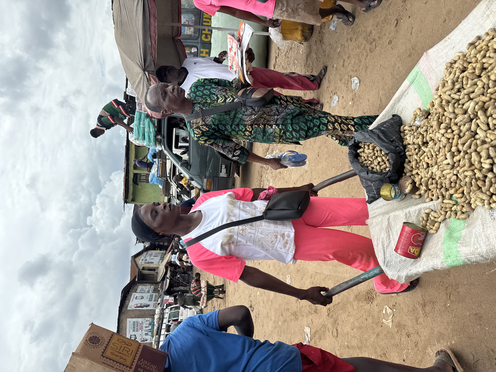 The image depicts a bustling market scene in Onitsha, Nigeria. A woman in pink pants and a white shirt with pink sleeves is pushing a wheelbarrow overflowing with peanuts. There are several cans and a black bag near the peanuts. A man in a patterned green shirt is holding a pair of sandals. Another man, partially visible at the bottom of the frame, wears a blue shirt and red shorts, and a box with "SIRI Forever Young" is visible. The background shows a variety of market stalls, buildings with advertising, and other people going about their business. The weather appears cloudy.