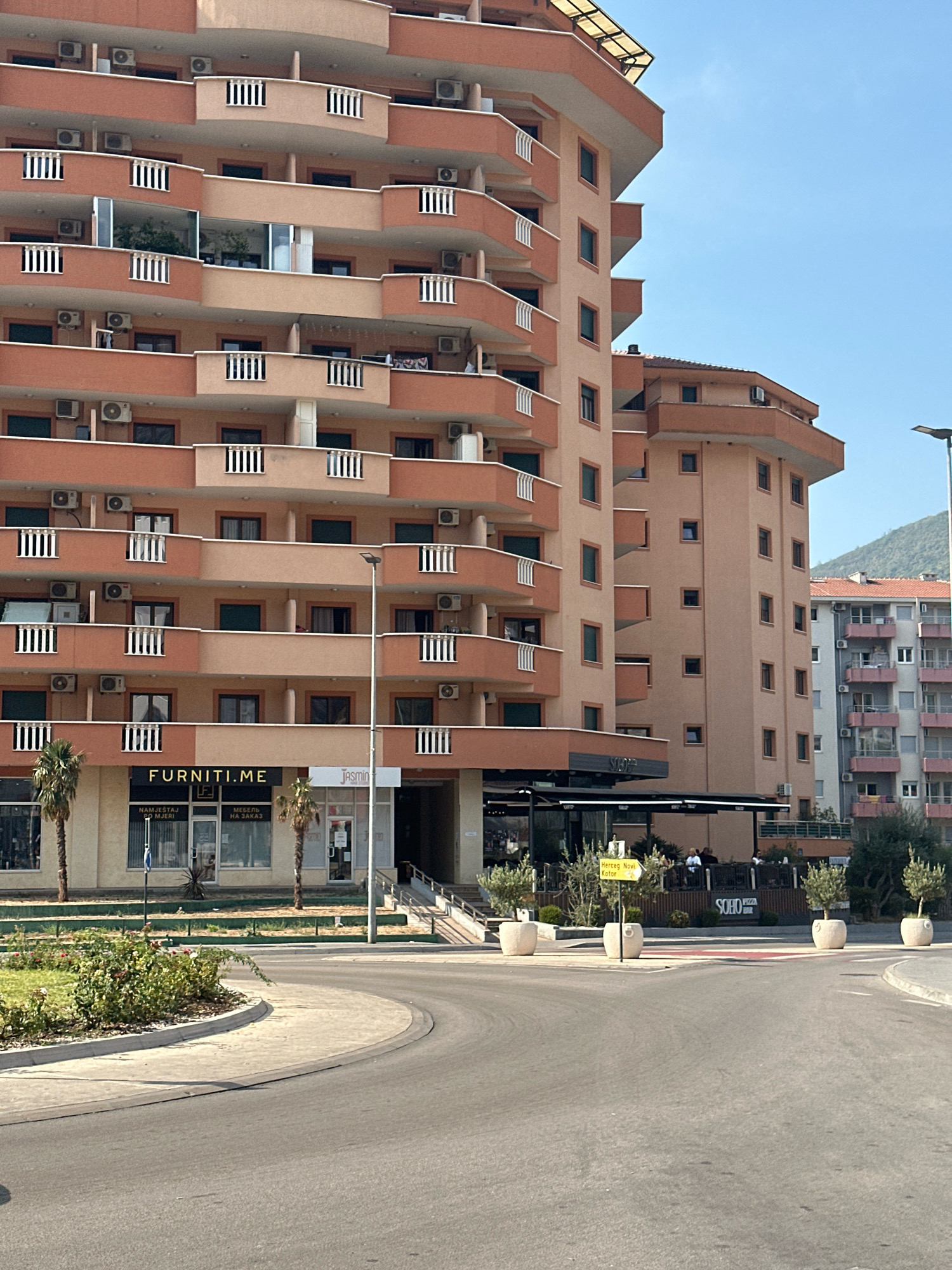 This is a medium-distance, eye-level shot of a street scene in Budva, Montenegro, focusing on a modern, multi-story apartment building with an attached commercial ground floor. The building is painted in shades of orange and beige, featuring numerous balconies with white railings. Air conditioning units are visible on many of the balconies. The ground floor houses commercial establishments, including a store with a sign reading "FURNITI.ME" and another with "JASMIN". A cafe or restaurant with outdoor seating is also present, indicated by tables and chairs under an awning, and a sign that says "SOHO PIZZA". Two palm trees stand in front of the building. A street sign points towards "Herceg Novi Kotor". The foreground shows a paved road with a curved intersection, a manicured lawn with some flowering bushes, and large, light-colored planters with small trees. In the background, another apartment building and a hillside with vegetation are visible under a clear blue sky. Several people are seated at the outdoor cafe.