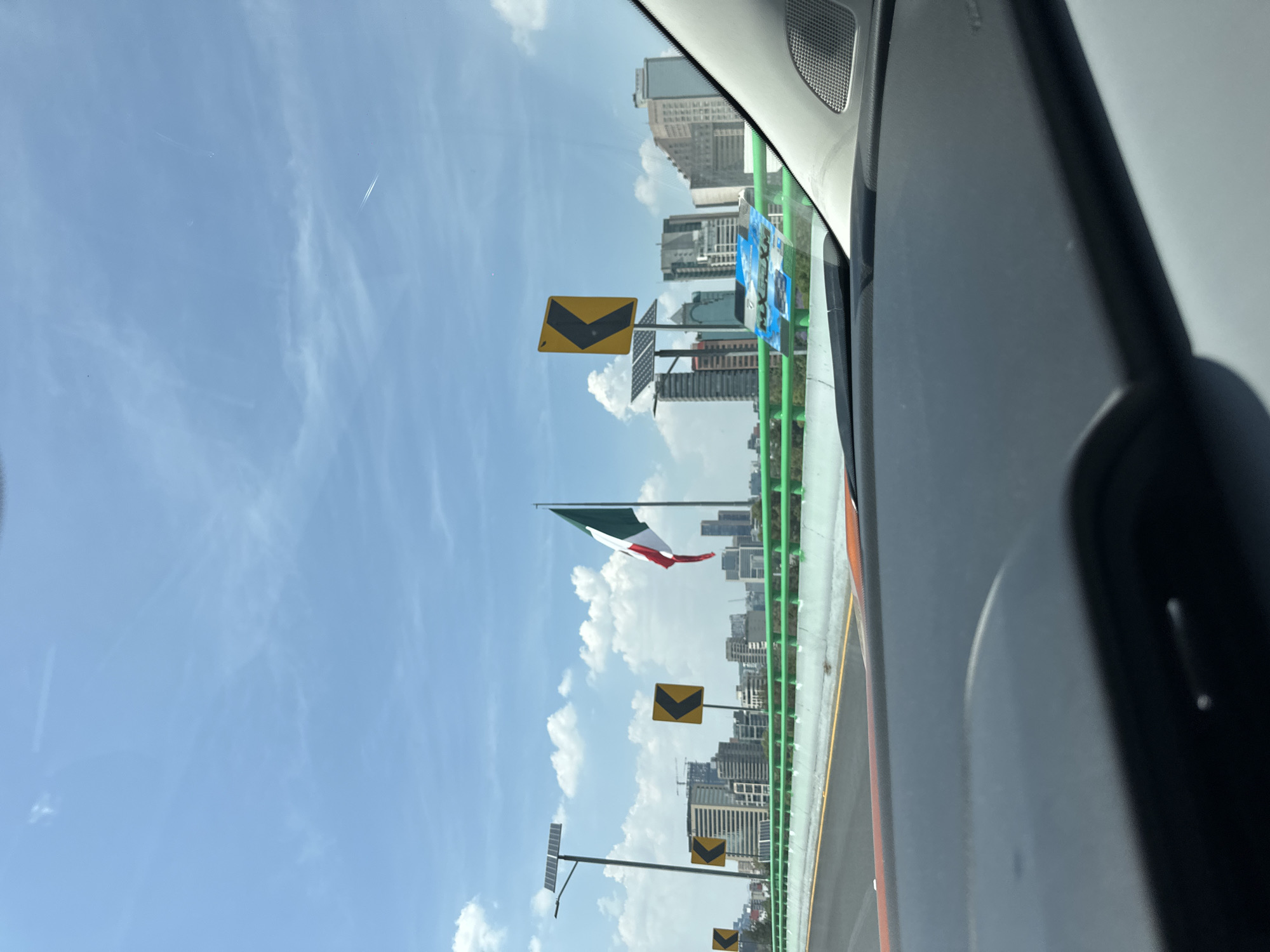 The image, taken from inside a vehicle, shows the exterior view of a bridge in Mexico City, Mexico. The sky is blue with scattered clouds. The Mexican flag is prominently displayed along the bridge. In the background are buildings, indicating an urban setting. The bridge has orange and green barriers. There are yellow road signs with a black chevron pointing downwards. The vehicle's interior is partially visible, including the dashboard.