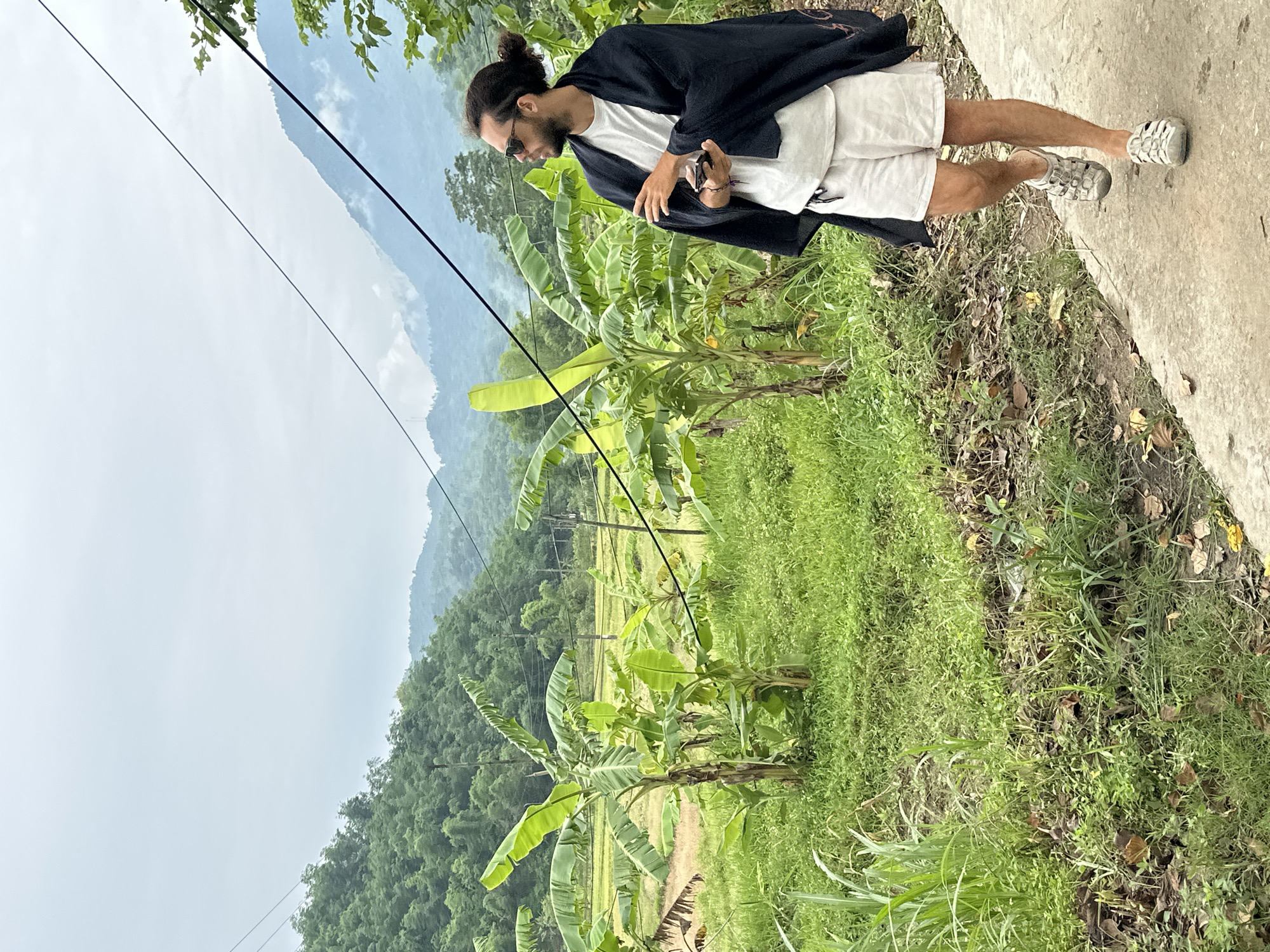 A man with dark hair pulled into a bun and wearing sunglasses, a white t-shirt, white shorts, and a dark shawl walks on a concrete path. He holds a dark-colored mobile phone in his right hand. The path runs alongside a lush green area with banana trees, grass, and other vegetation. In the background, terraced rice paddies are visible, and further beyond, rolling hills and mountains covered in trees rise under a cloudy sky. The scene is located in Mai Chau, Vietnam.