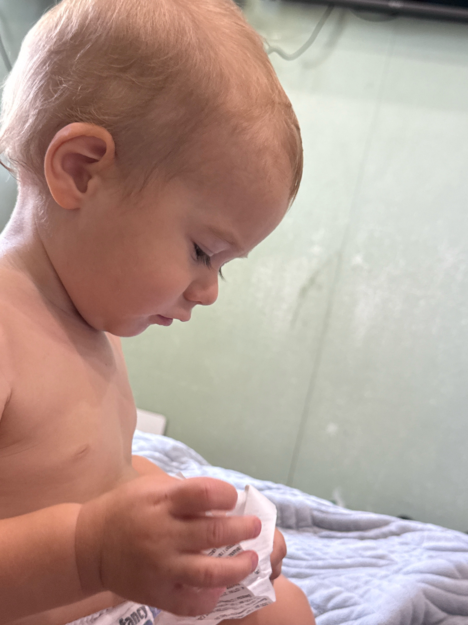 An infant, viewed from the chest up in a profile shot, is seated on a blue patterned blanket. The infant's head is tilted downwards, with their gaze focused on a small, crumpled white object held in their hands. The infant has fair skin and light brown hair, and their bare chest is visible. The background is a plain, light green wall. There are no identifiable landmarks indicating the city of Pavlodar, Kazakhstan.