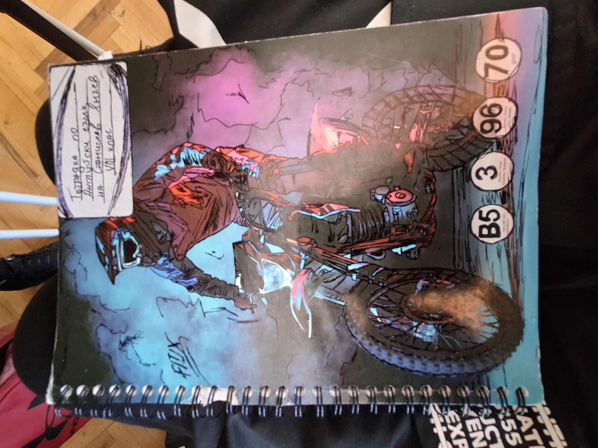 The image shows a notebook with a graphic of a motocross rider on a motorcycle. A handwritten label is affixed to the top right corner of the notebook cover. The label is written in Cyrillic script and appears to identify the notebook's subject and owner. The overall scene is dimly lit, with the notebook resting on dark fabric, possibly clothing or a bag. The background is out of focus, revealing wooden flooring and some light-colored objects. The presence of a notebook with a label in Cyrillic suggests it belongs to a student in a Bulgarian-speaking region. While Varna, Bulgaria, is mentioned, there are no specific visual cues in the image to confirm its location within that city.