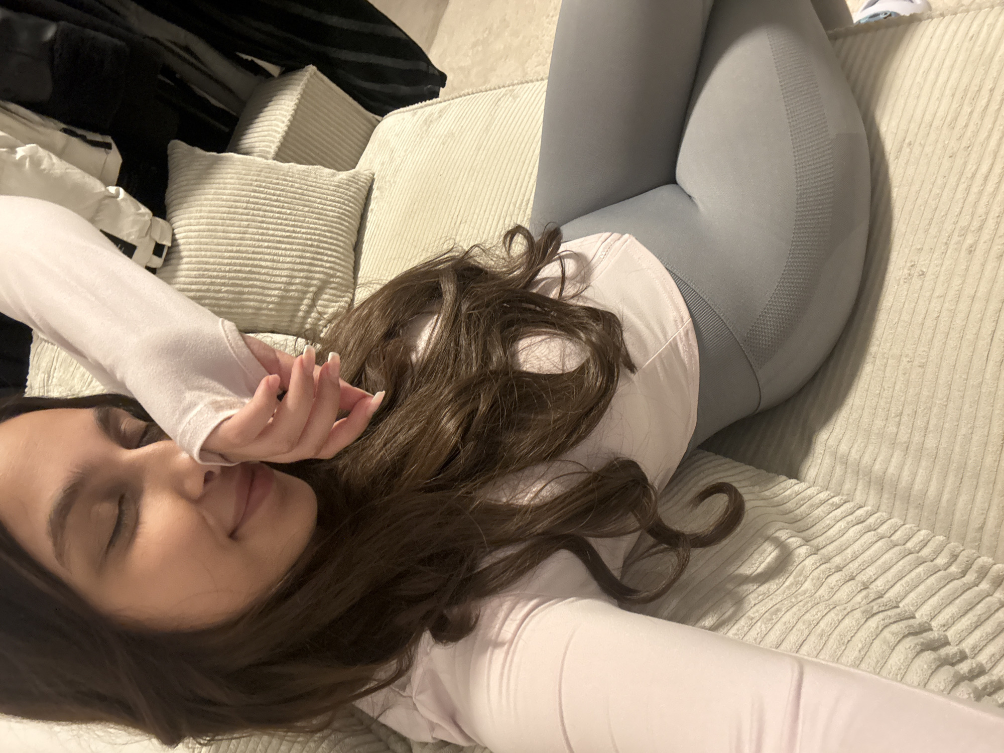 A woman is lying on a corduroy couch. She is wearing grey leggings and a white long-sleeved shirt. Her dark hair is spread around her head. Her eyes are closed, and she is smiling slightly. Her hand is near her face. The couch is in a room with pillows and a dark object behind it.
This image cannot be used to identify any location in Mönchengladbach, Germany.