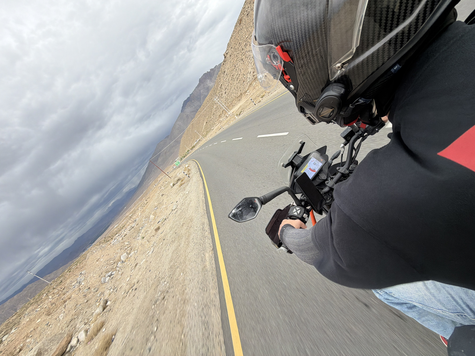 A first-person perspective shot shows a person riding a motorcycle on a paved road with a yellow line and white dashes, winding through a desolate, mountainous landscape. The rider wears a black long-sleeved shirt, blue jeans, and a grey sleeve on their left arm. A carbon fiber patterned helmet is on their head, with a clear visor. The motorcycle has a digital display on the handlebars, and a small camera is mounted on the tank. The environment is arid, with tan-colored hills and sparse vegetation. The sky is overcast with dramatic, dark clouds. In the distance, mountains are visible. The road appears to be in the vicinity of Chumkar, India, based on the terrain and type of road.